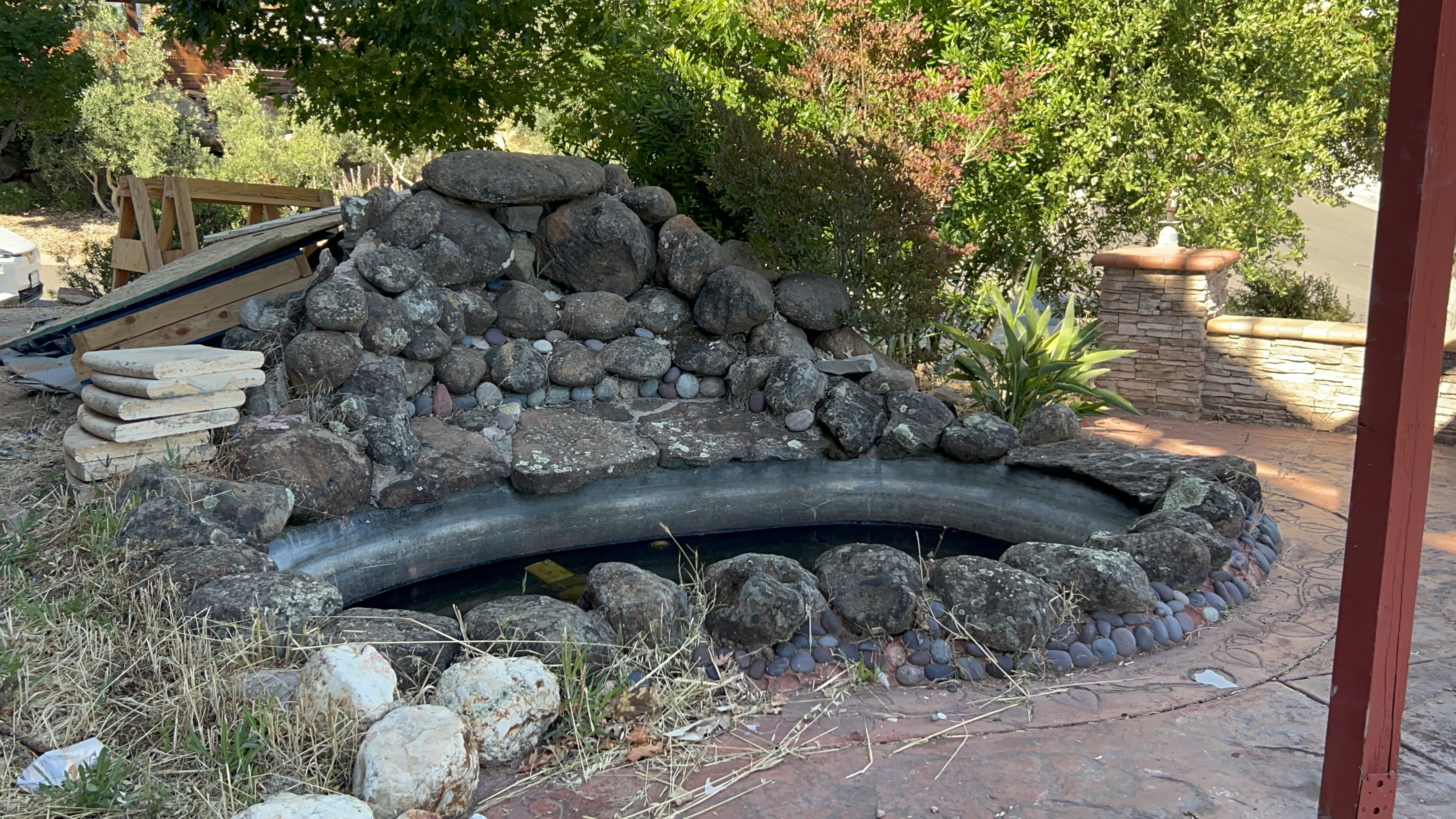 This outdoor space features a custom-built, kidney-shaped rock pond integrated into a landscaped garden setting. The water feature is framed by natural stone boulders and decorative river pebbles, set against a backdrop of lush green foliage and a stone-clad pillar. It creates a rustic, tranquil focal point suitable for a garden or backyard retreat.