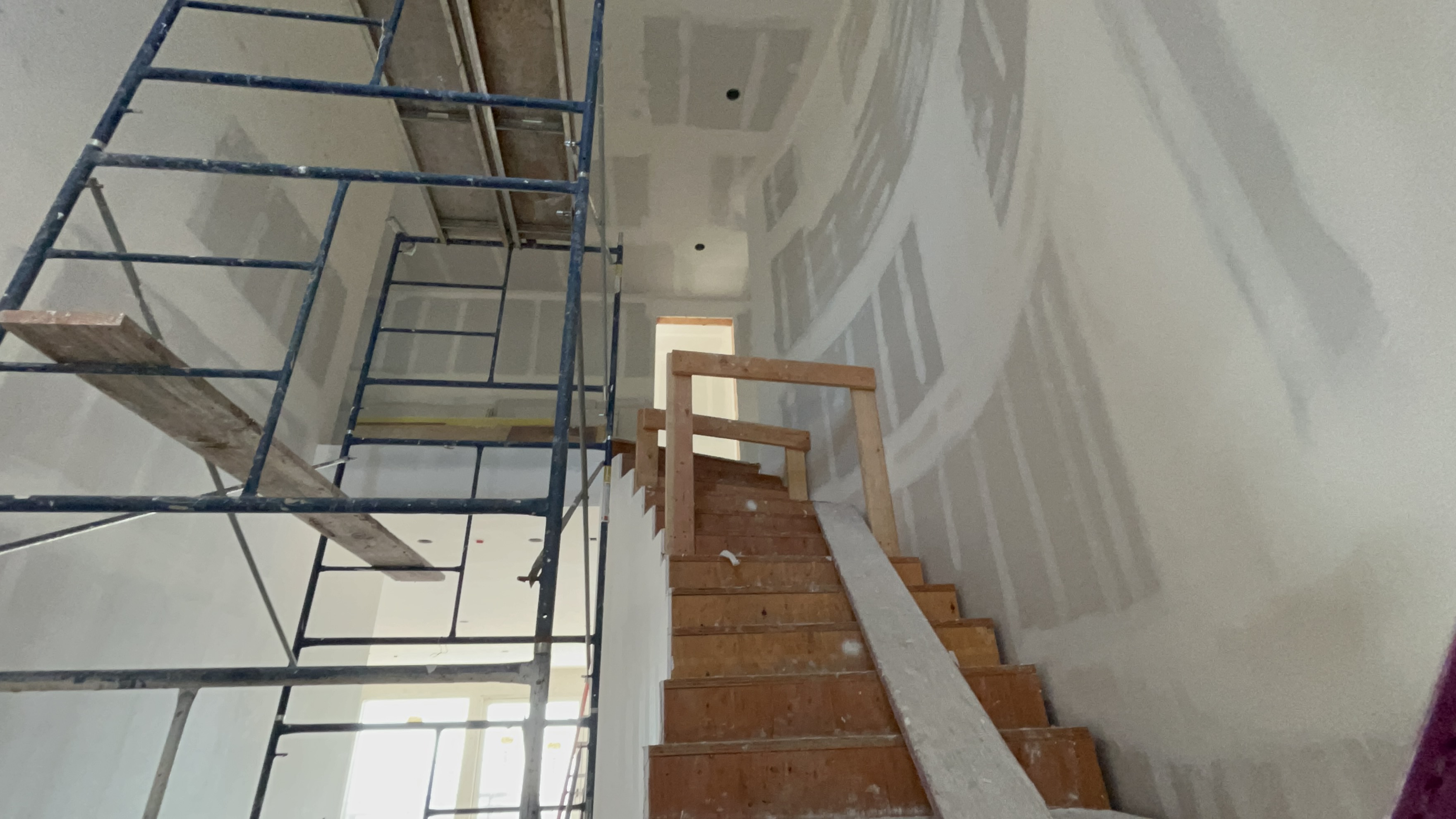This image shows a staircase under construction within an unfinished home. The space features temporary scaffolding, exposed drywall with visible mudding, and unsealed wooden stair treads, reflecting an ongoing renovation or new build project. It offers a glimpse of the structural layout and ongoing development of the property's entryway or central corridor.