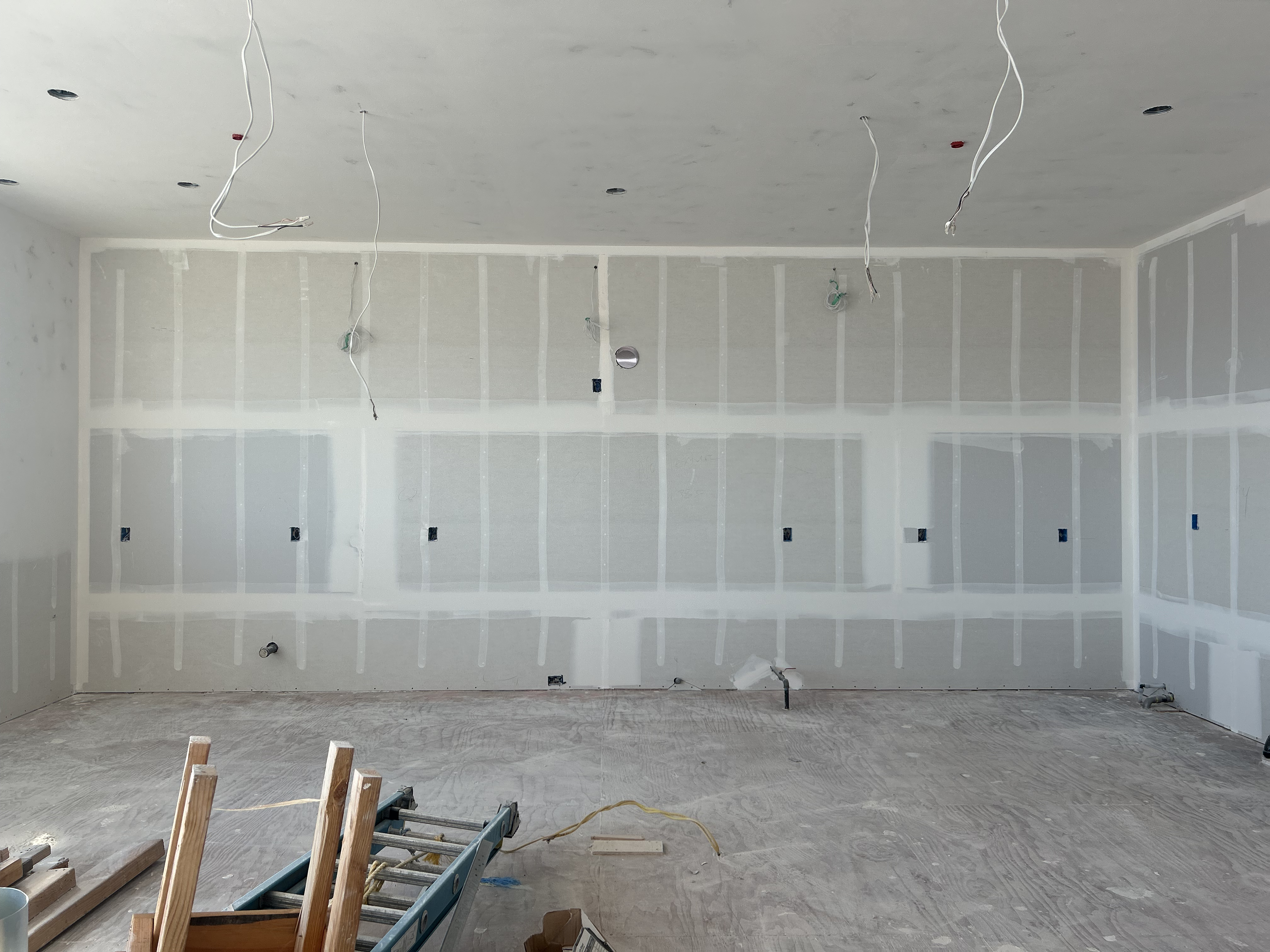 This image shows a room under construction, likely a kitchen, characterized by unfinished drywall walls and exposed electrical wiring hanging from the ceiling. The layout includes multiple electrical outlet boxes and plumbing rough-ins, suggesting a space prepared for cabinets, appliances, and fixtures. Its current state represents an open-concept project ready for custom finishes and personalization.