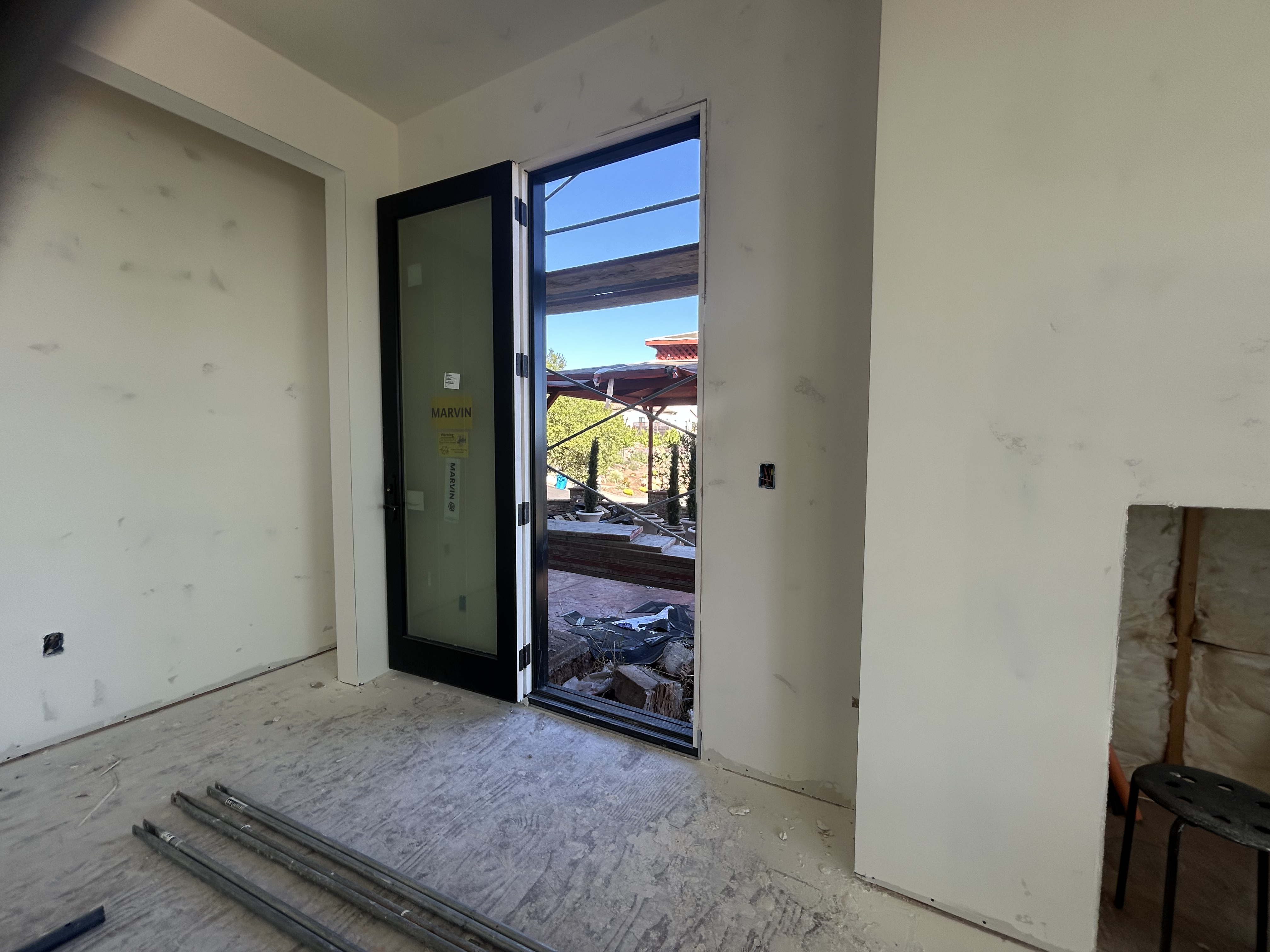 This image shows a room under construction, featuring an open doorway with a modern black-framed Marvin glass door leading to an outdoor area. The walls have been drywalled and are awaiting final finishes, while the floor consists of unfinished subflooring. To the right, a recessed area exposes wall insulation, indicating an ongoing renovation or new construction project.