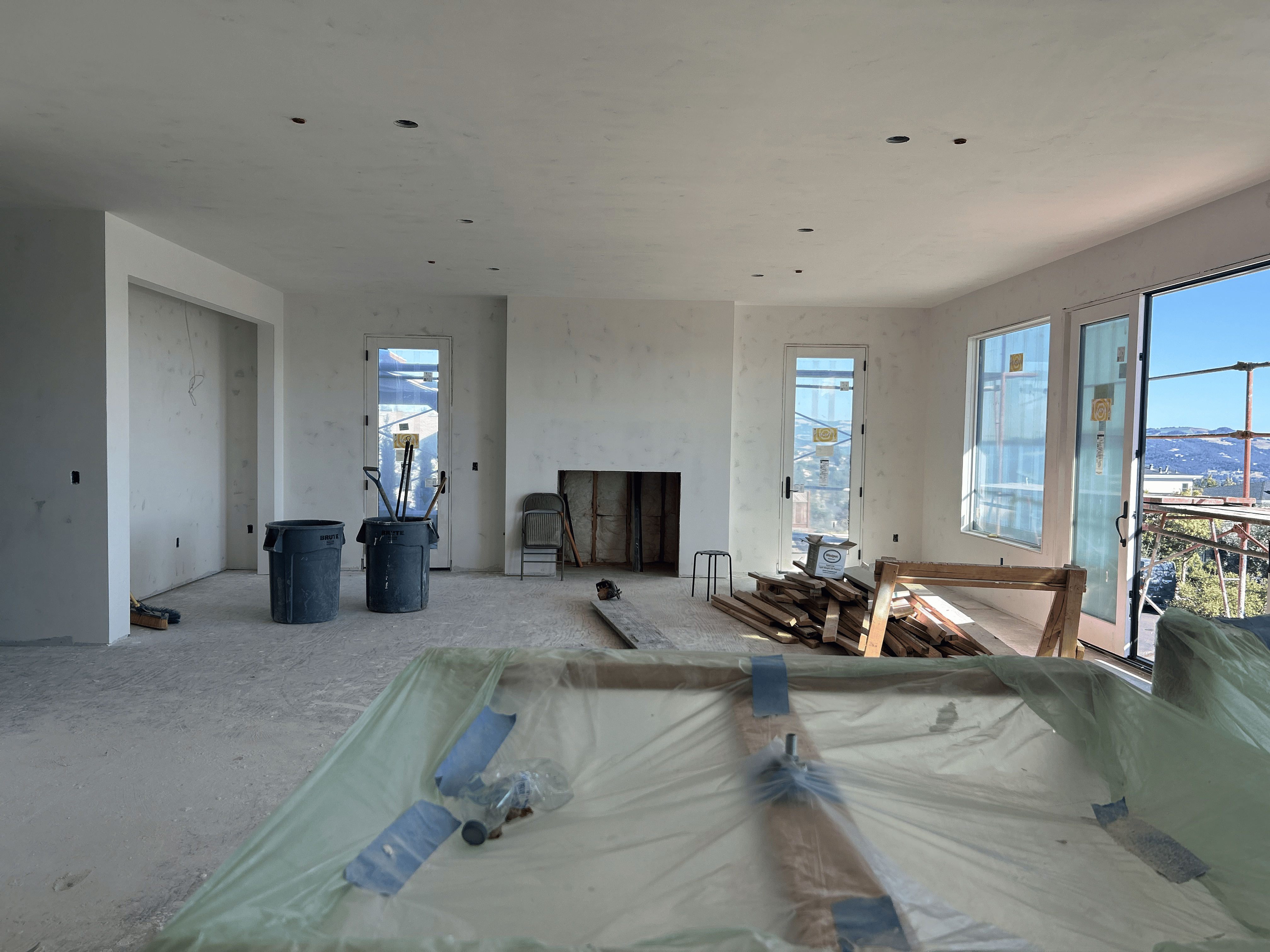 This image shows a spacious, unfinished living room area currently under construction, featuring white walls, neutral flooring, and a large fireplace opening centrally located on the back wall. The space is brightened by several glass doors and large windows that offer expansive views of the surrounding hills, suggesting a modern open-concept design potential. Construction materials like wood scraps, waste bins, and tools are visible, indicating the project is in the final stages of build-out and awaiting finishing touches.