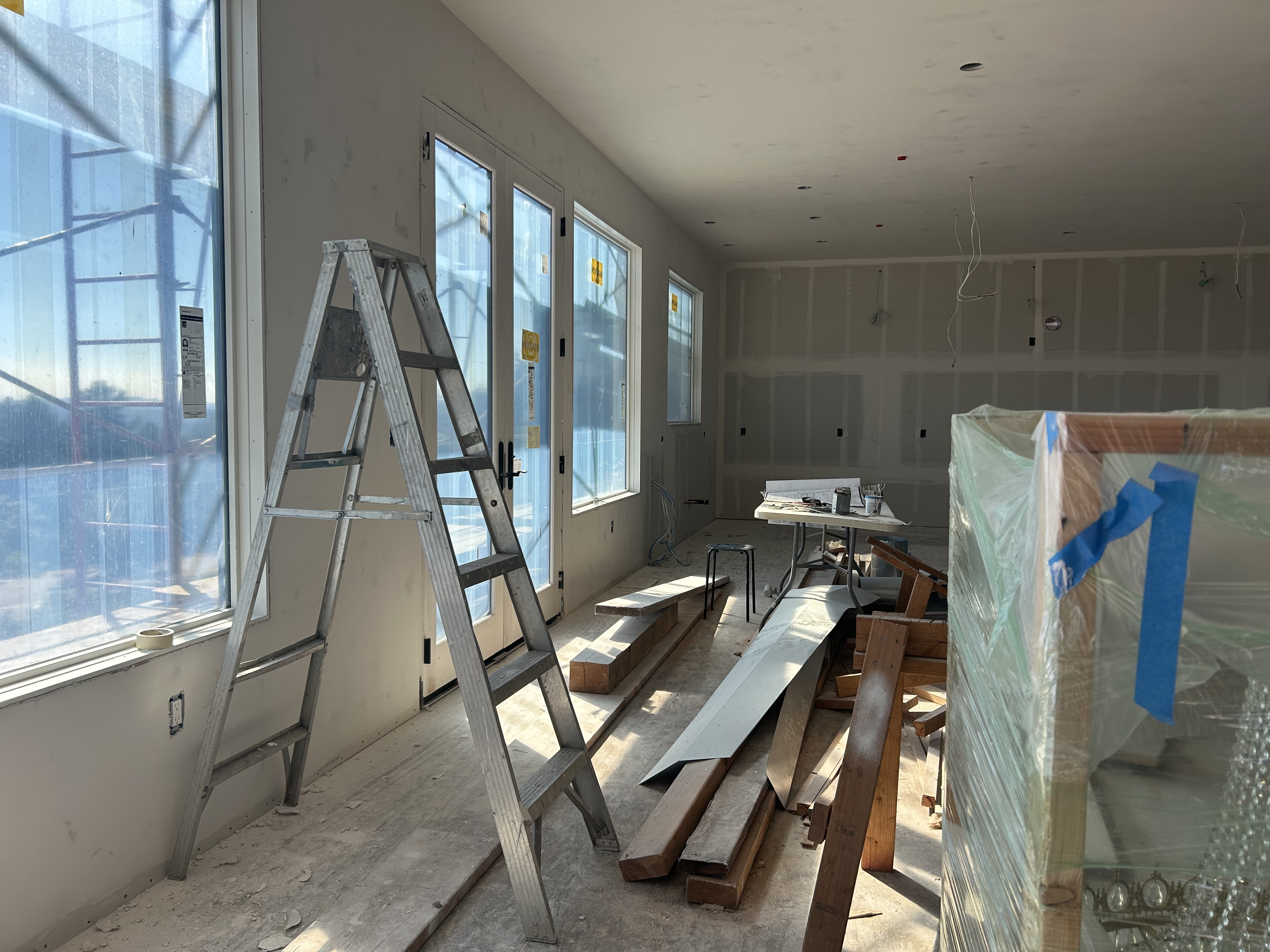 This image shows a room under construction, featuring unfinished drywall, exposed wiring, and construction debris like ladders and wood planks scattered throughout. Large floor-to-ceiling windows and a set of exterior doors allow for ample natural light, suggesting an open-concept living or dining space currently being finished. The presence of a wrapped furniture item indicates ongoing interior design or furnishing work in a newly built or renovated residential space.