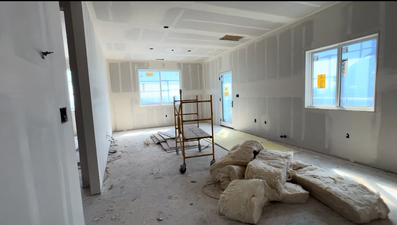 This interior space is currently under construction, featuring bare drywall, exposed subflooring, and scattered building materials. The room includes multiple windows providing natural light, a neutral layout, and an open floor plan structure. It offers a blank canvas for potential buyers looking to customize a new home build.