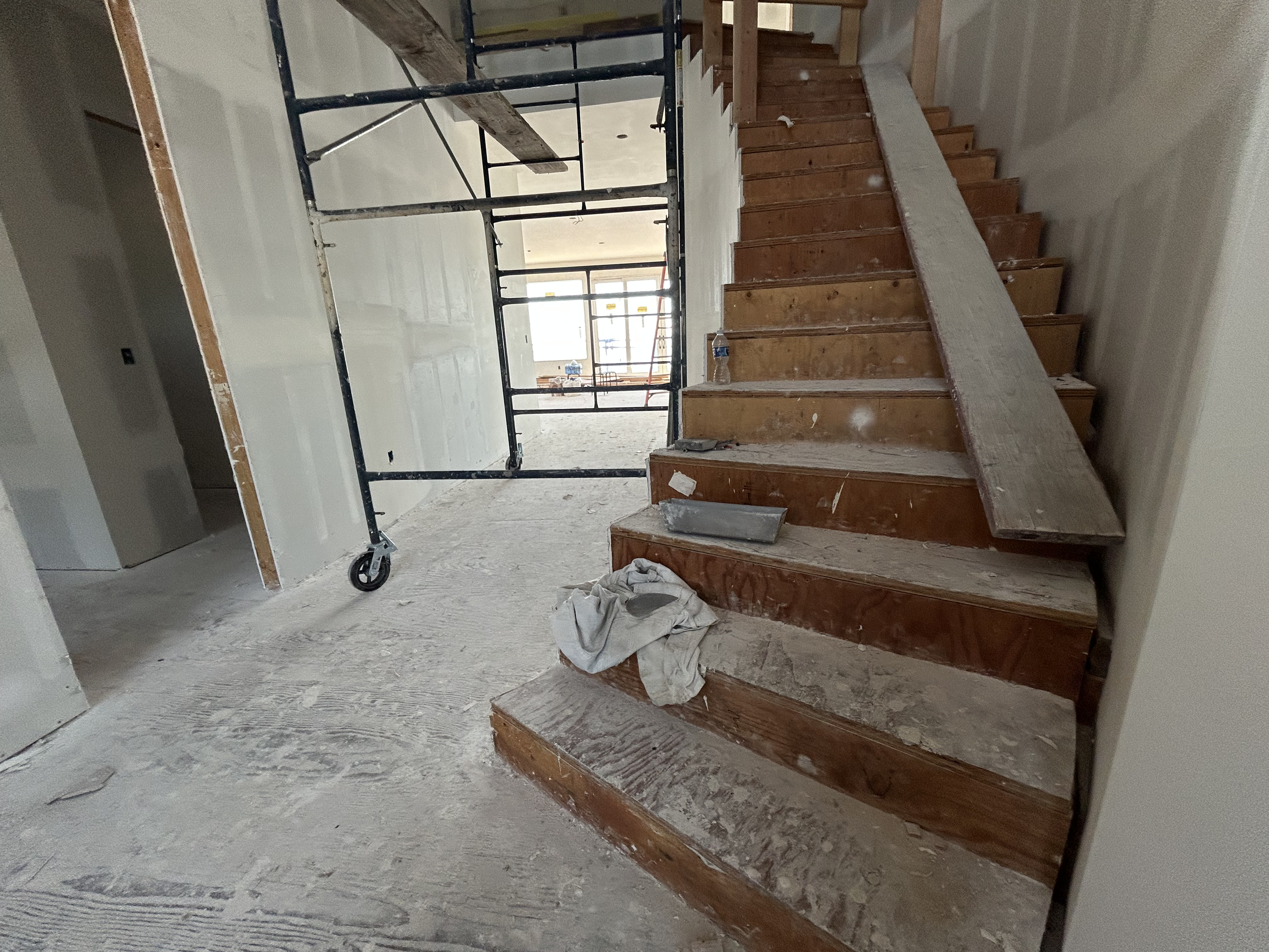 This image shows a home currently under construction, featuring an unfinished wooden staircase at the center of the frame. In the background, scaffolding stands in a hallway that leads toward an open, sunlit living area. The interior showcases raw drywall and exposed materials, highlighting the potential for a customizable and modern design.