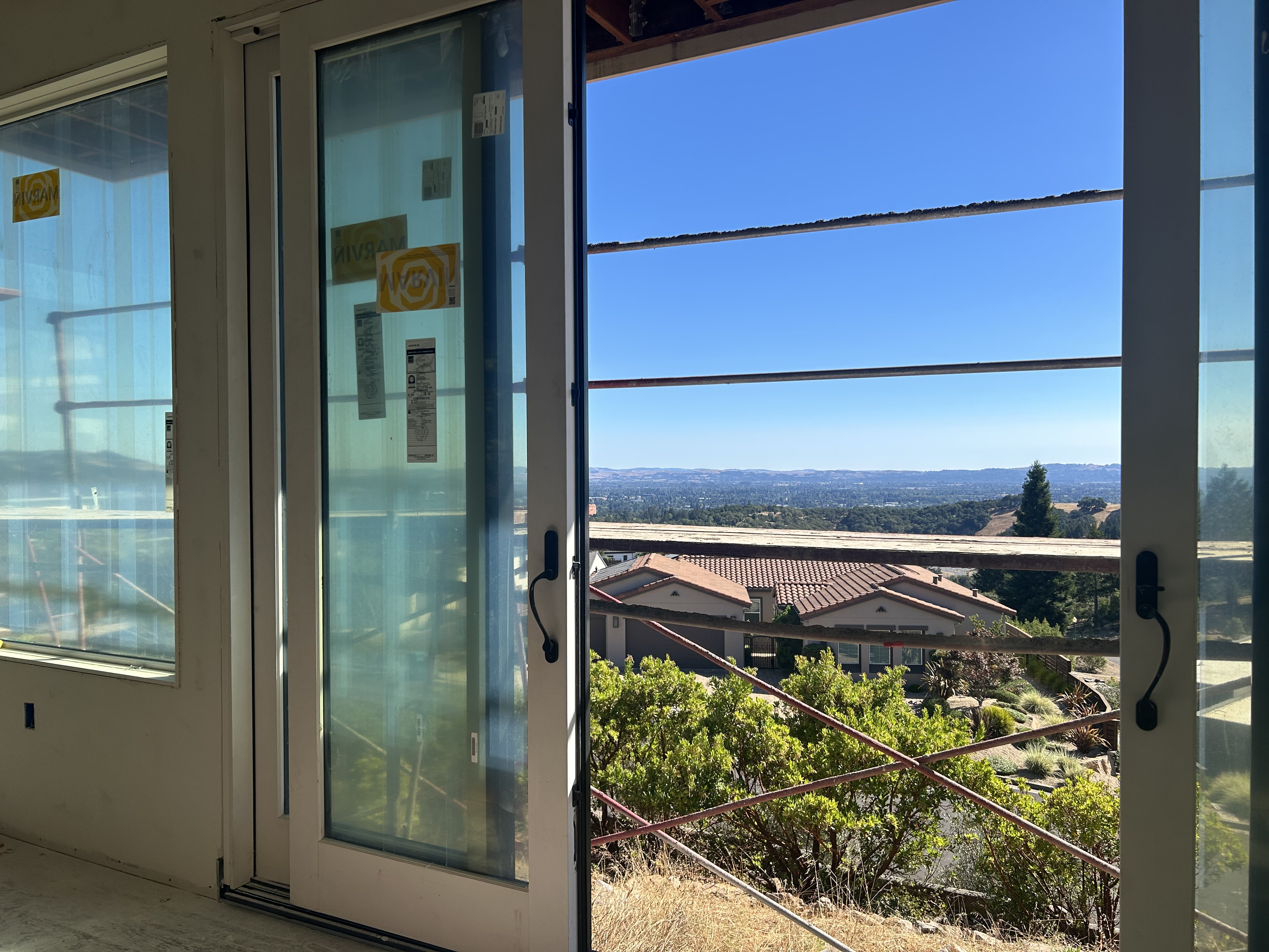 This image shows a room under construction, featuring large sliding glass doors that open up to an expansive, elevated panoramic view of a residential valley and hills. The sliding door system includes a prominent frame with manufacturer labels still attached, suggesting a new installation in a home focused on indoor-outdoor living. Scaffolding is visible outside, indicating ongoing renovation or construction work on the property.