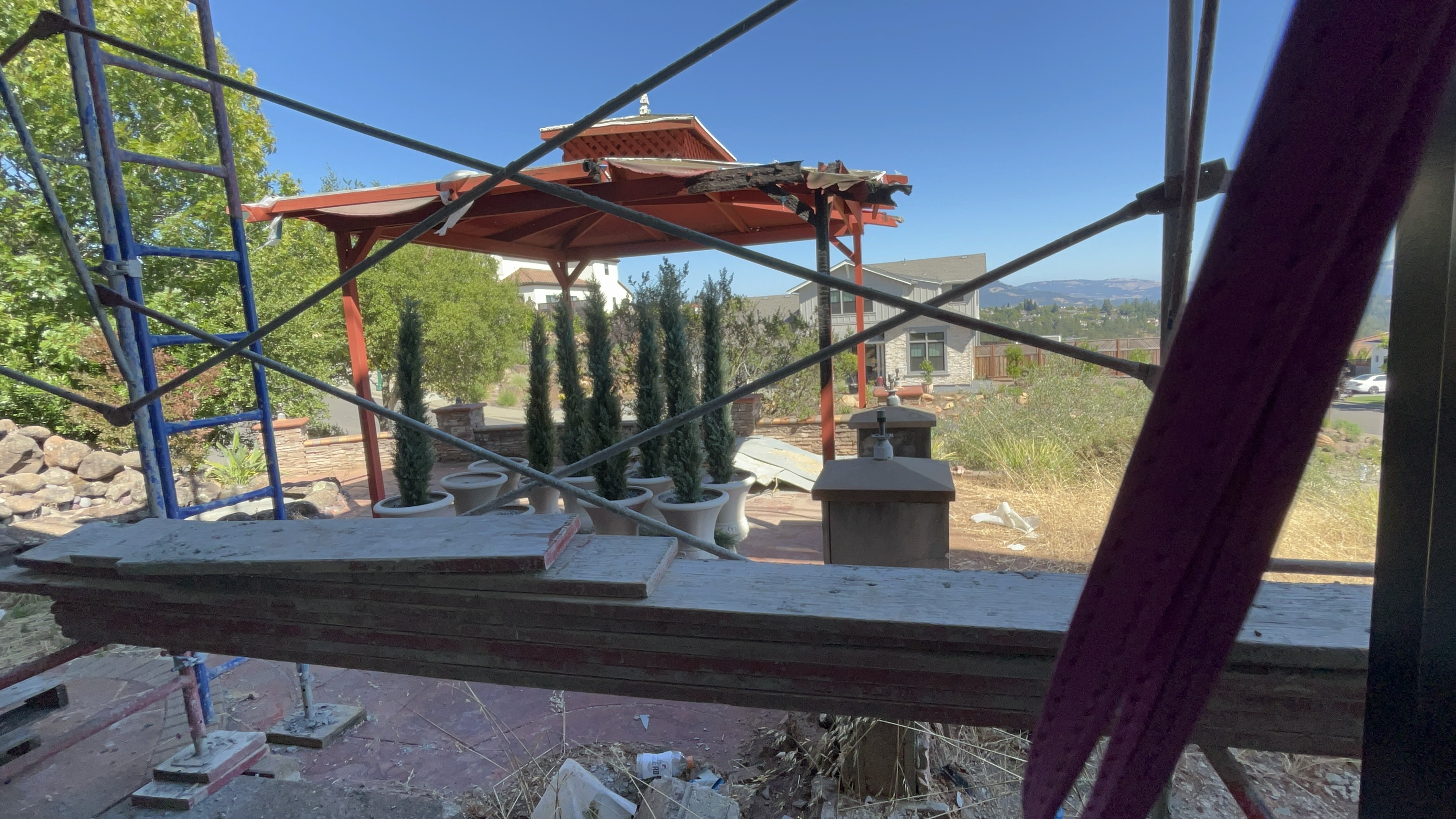 This outdoor space features a partially damaged gazebo structure under repair, supported by scaffolding in the foreground. The patio area includes a series of potted evergreen shrubs set against a stone wall, with a view of a neighboring home and distant hills in the background. While currently undergoing renovation, the area offers potential for a pleasant, elevated outdoor lounge space.