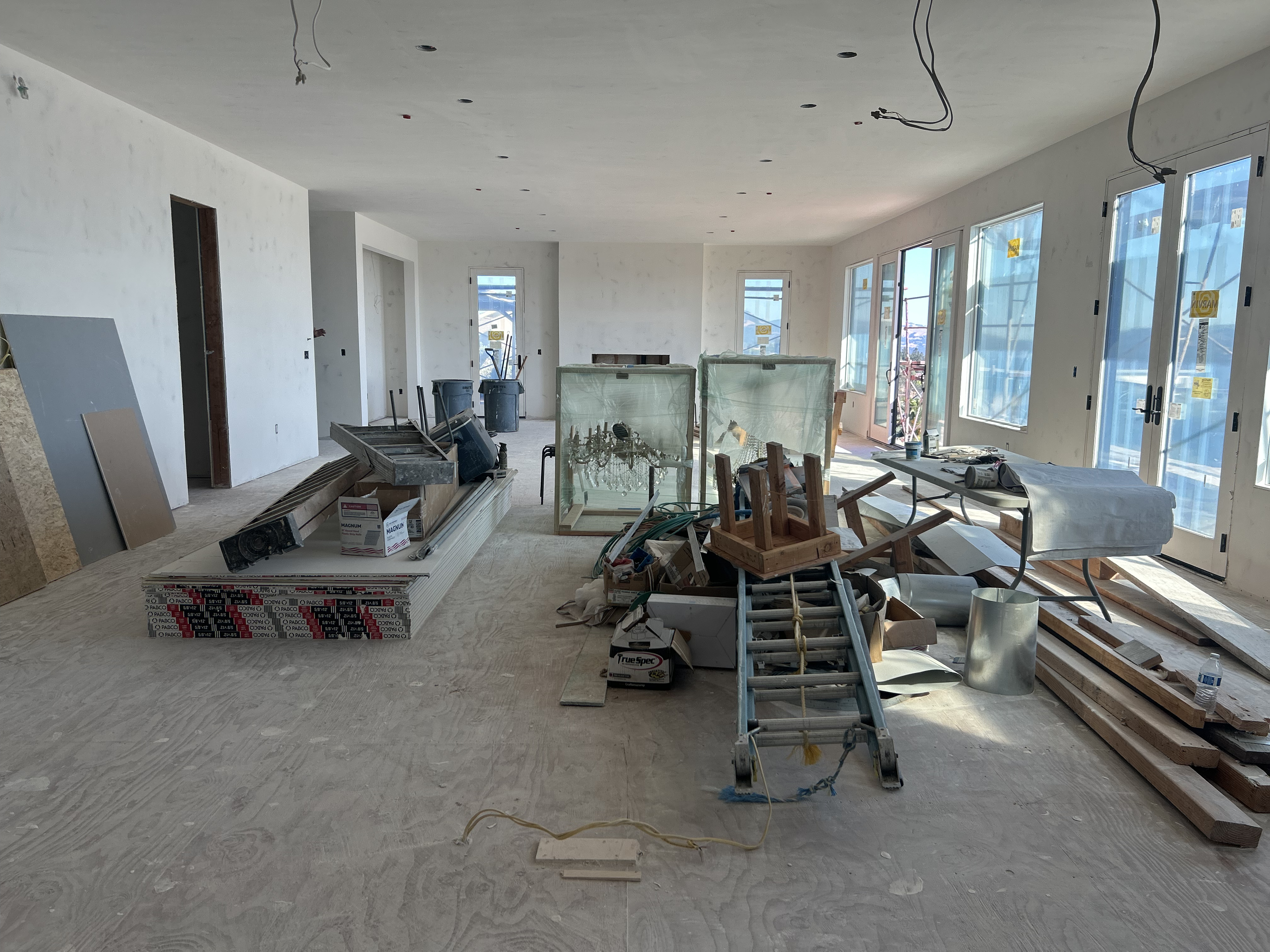 This spacious, unfinished room is currently under construction, featuring bare drywall and exposed wiring throughout. A large sliding glass door system along the side wall provides ample natural light and views, while a stack of construction materials and a chandelier case sit in the center of the room. It offers a generous, open-concept layout suitable for a potential living area once fully completed.