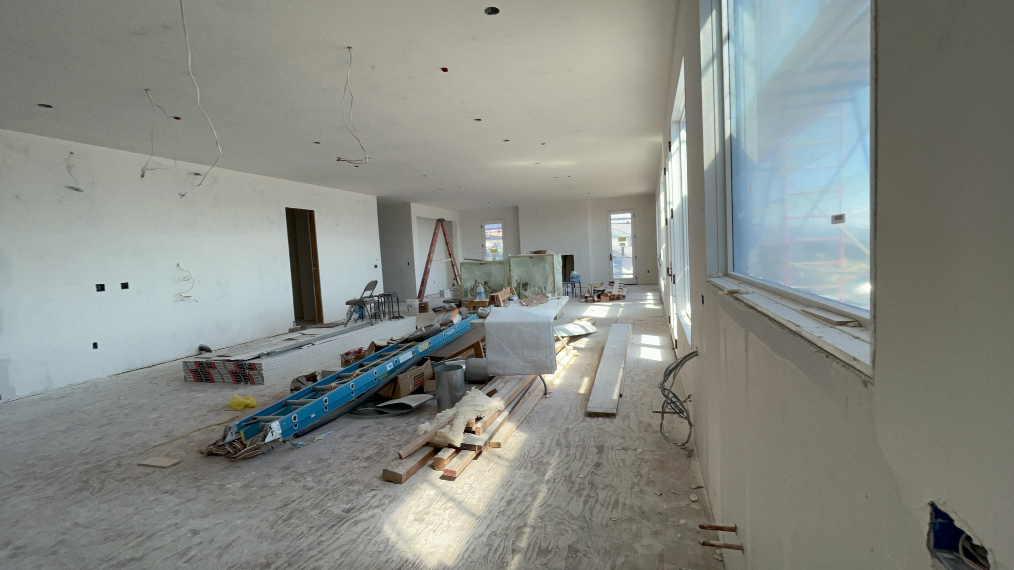 This image shows a spacious, unfinished interior room currently under construction, featuring bare walls, plywood flooring, and exposed wiring. A large bank of windows along the right wall provides significant natural light, suggesting an open layout suitable for a living or multi-purpose area. The space is filled with construction equipment, tools, and materials, indicating an ongoing renovation or new build project.