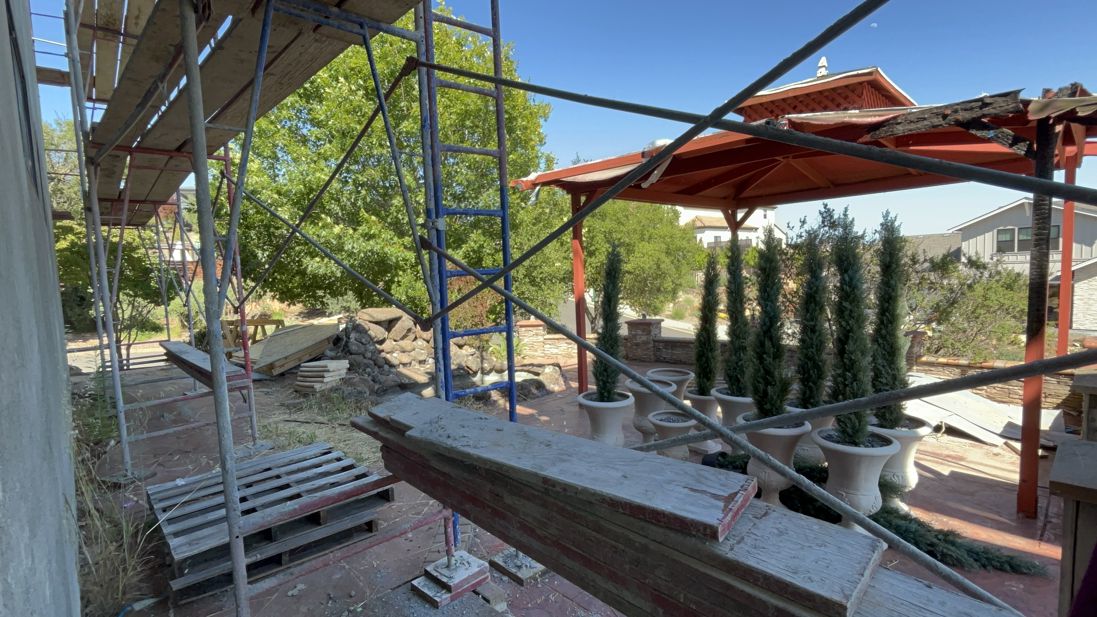 This outdoor patio area is currently undergoing construction, featuring scaffolding and stacked building materials set against an orange-red pergola. Several potted evergreen topiary trees are arranged in a row, adding a touch of structure to the space. The area suggests a renovation project focused on enhancing an outdoor entertaining or garden space.