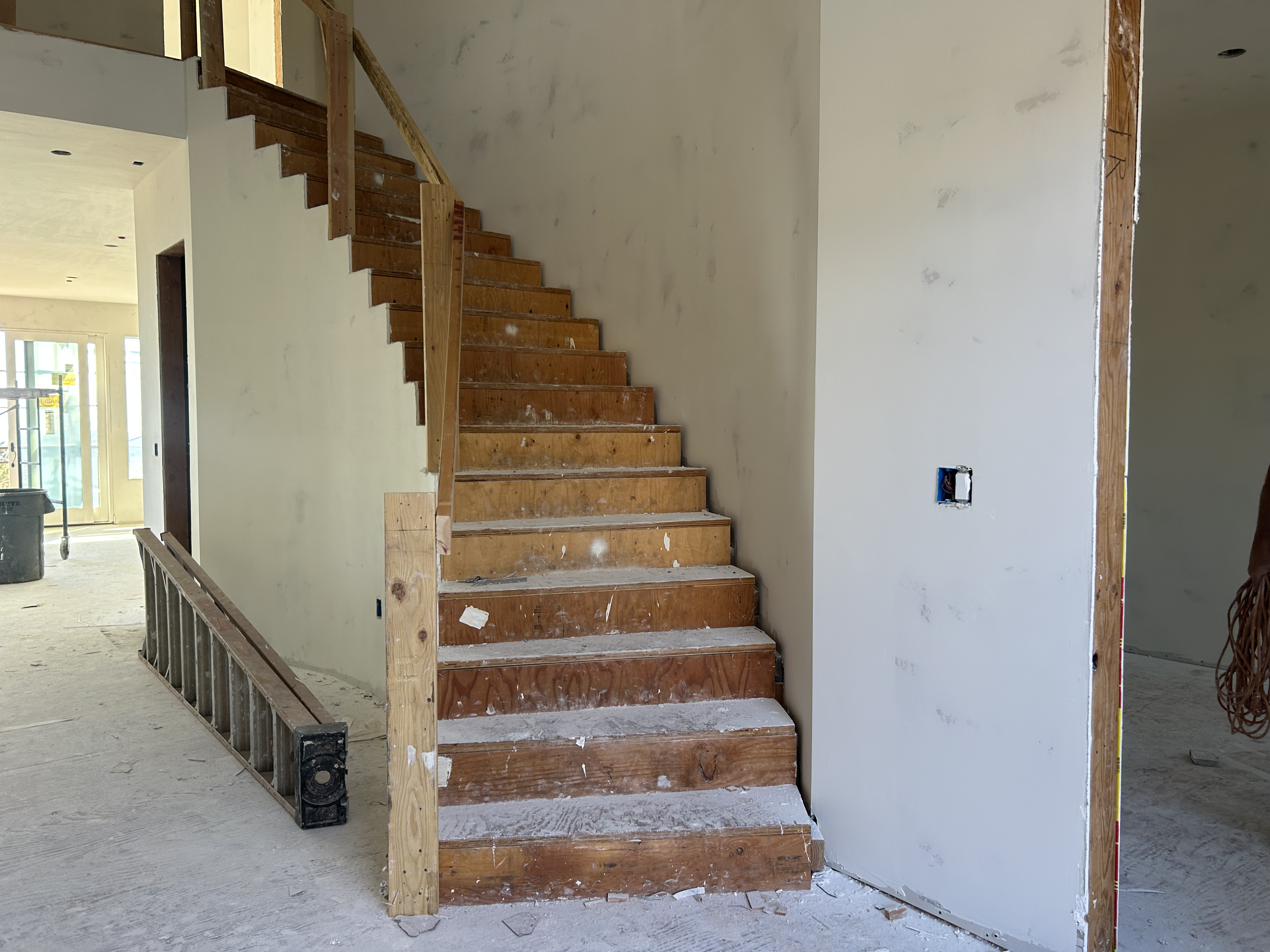 This image shows a staircase currently under construction within a new home build. The unfinished wooden stairs and drywall reveal a project in progress, offering prospective buyers a glimpse into the home's structure and layout. The space features high ceilings and a neutral, primed canvas ready for customized finishes like carpet, hardwood, or decorative trim.