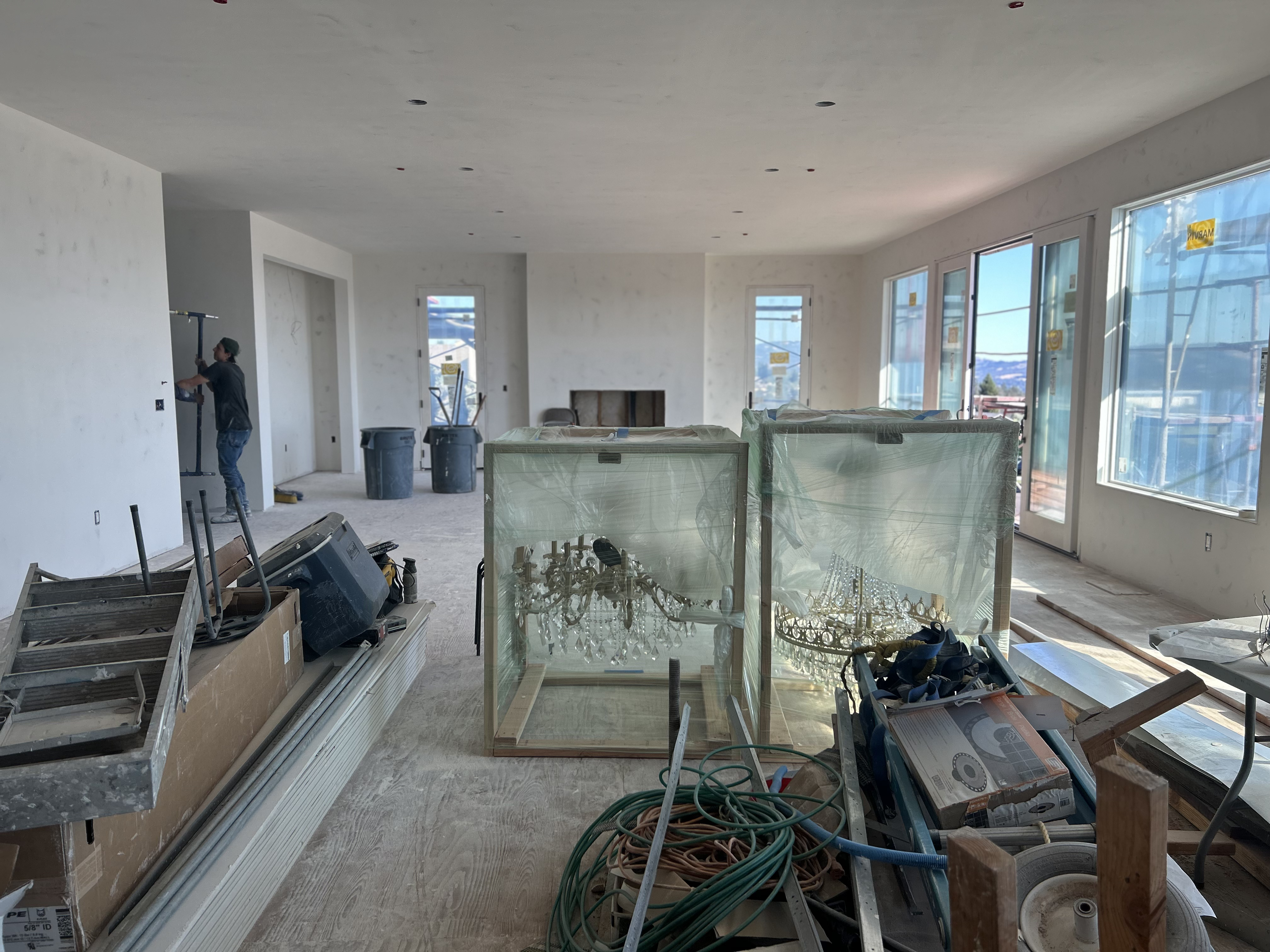 This interior space currently under construction reveals a spacious, open-concept living area with large floor-to-ceiling windows that provide ample natural light and scenic views. The room features high, smooth-finished ceilings and is awaiting final touches, with large boxed chandeliers prepped for installation. The expansive layout highlights the potential for a bright, modern living space suitable for a luxury home.