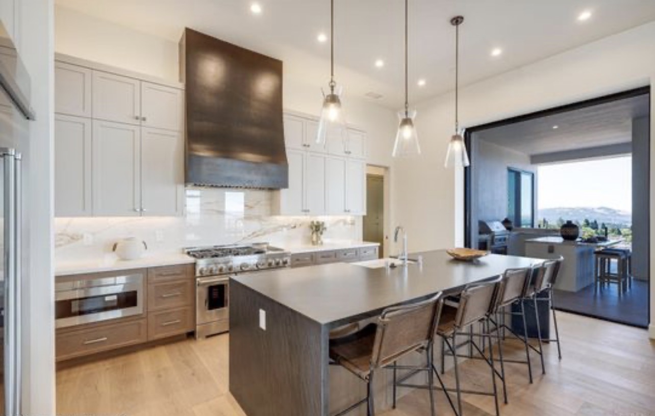 This sleek, modern kitchen features a two-tone cabinetry design with light upper cabinets and rich-toned wood lower cabinets, complemented by a large central island with bar seating. The space is anchored by an impressive dark-finished range hood and professional-grade stainless steel appliances, all set against elegant marble-look backsplashes. Large glass doors open to an outdoor space, creating a seamless connection between the kitchen and the scenic exterior views.