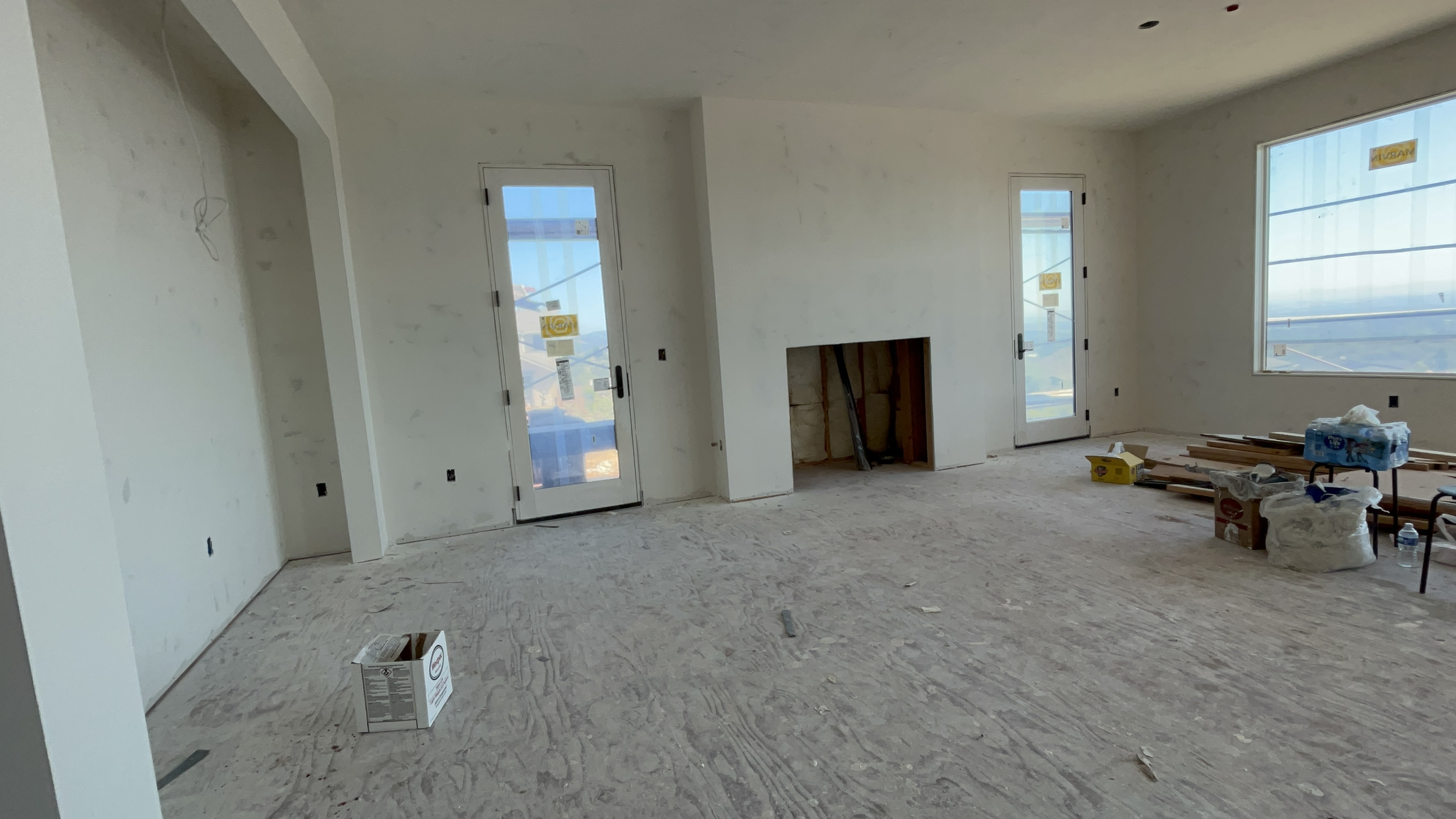 This living room is currently under construction, featuring unfinished drywall, plywood subflooring, and an open fireplace cutout. The space is illuminated by two glass-paned doors and a large window, which provide ample natural light and views of the surrounding landscape. It represents a blank canvas ready for custom finishes and flooring options.
