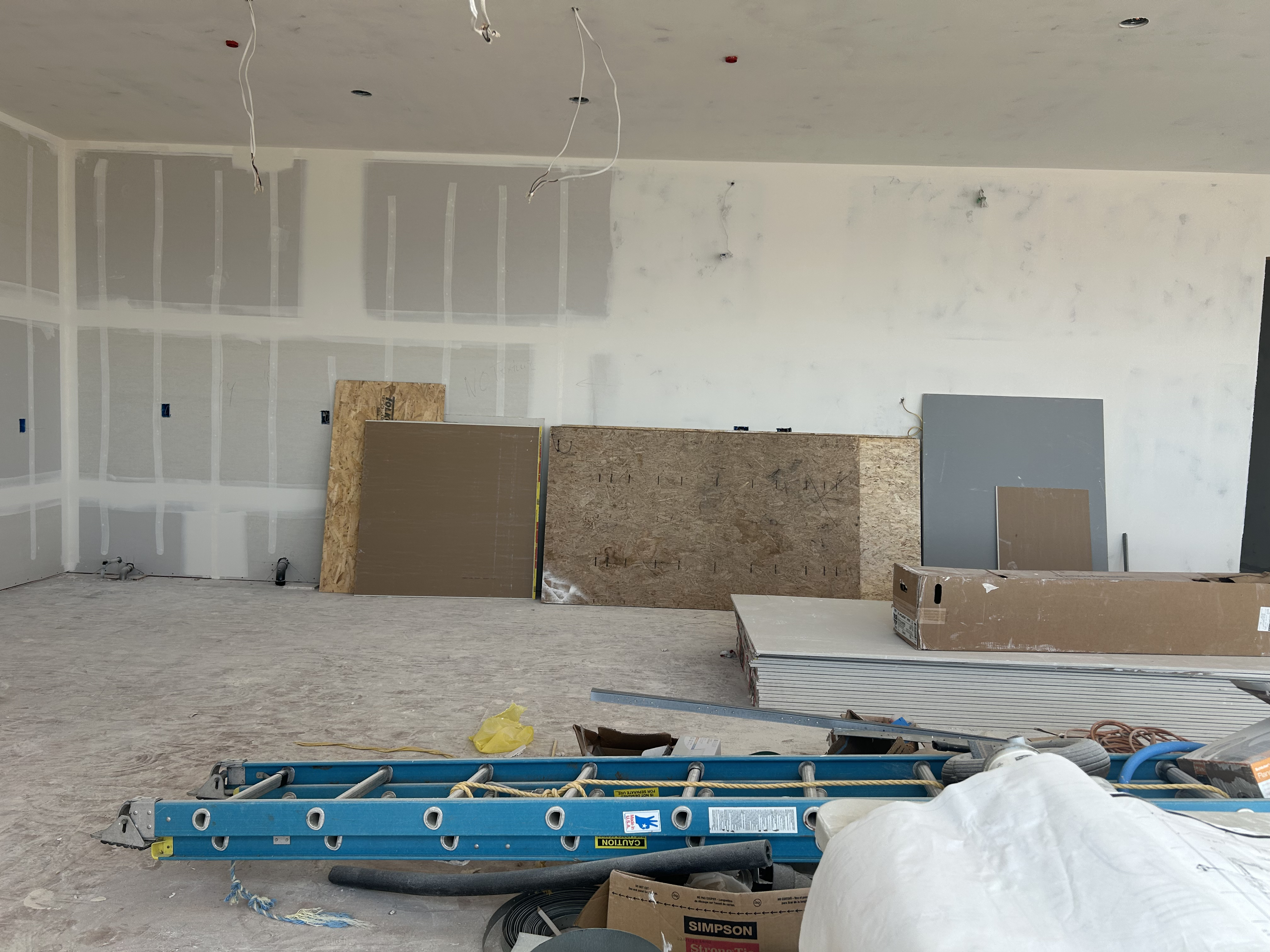 This image captures a room under construction, featuring bare, unfinished drywall with visible tape and joint compound. The floor is covered in construction debris, and various building materials, such as stacks of drywall, plywood, and an extension ladder, are scattered throughout the space. It is an unfinished interior area, likely a living or bonus room, currently undergoing renovations.