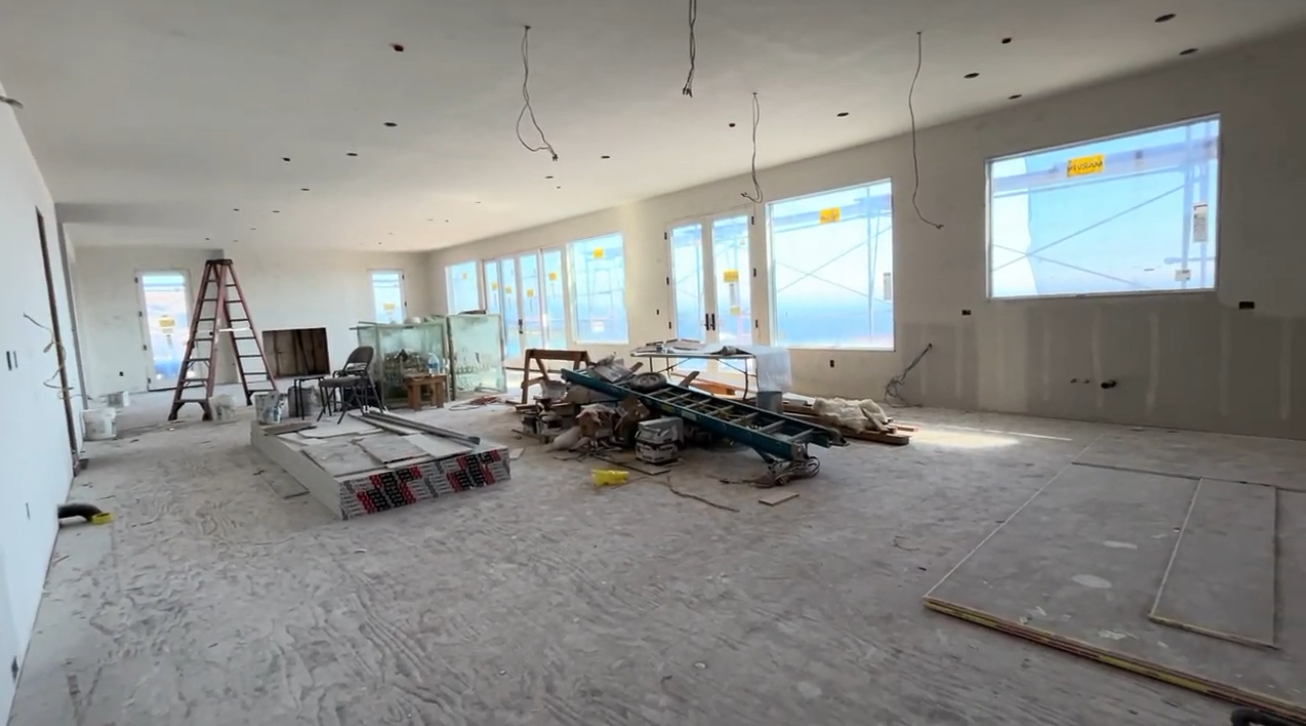 This is a large, open-concept living area currently under construction, featuring unfinished wood floors and pristine, unpainted drywall. The space is filled with natural light streaming through multiple large windows and glass doors, which offer a clear view of scaffolding outside. With its expansive layout and high ceilings, this room presents a blank canvas that could be transformed into a sophisticated and airy main living space.
