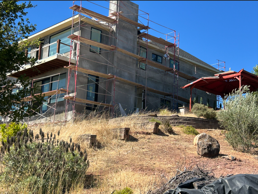 This image shows the rear exterior of a multi-story home currently undergoing extensive facade renovation, evidenced by the prominent scaffolding covering much of the structure. The house features a modern architectural design with large windows, an unfinished stucco finish, and a raised wooden balcony. The property is situated on a sloped lot with a tiered stone retaining wall and a red gazebo, suggesting significant potential for outdoor living spaces once landscaping and construction are completed.