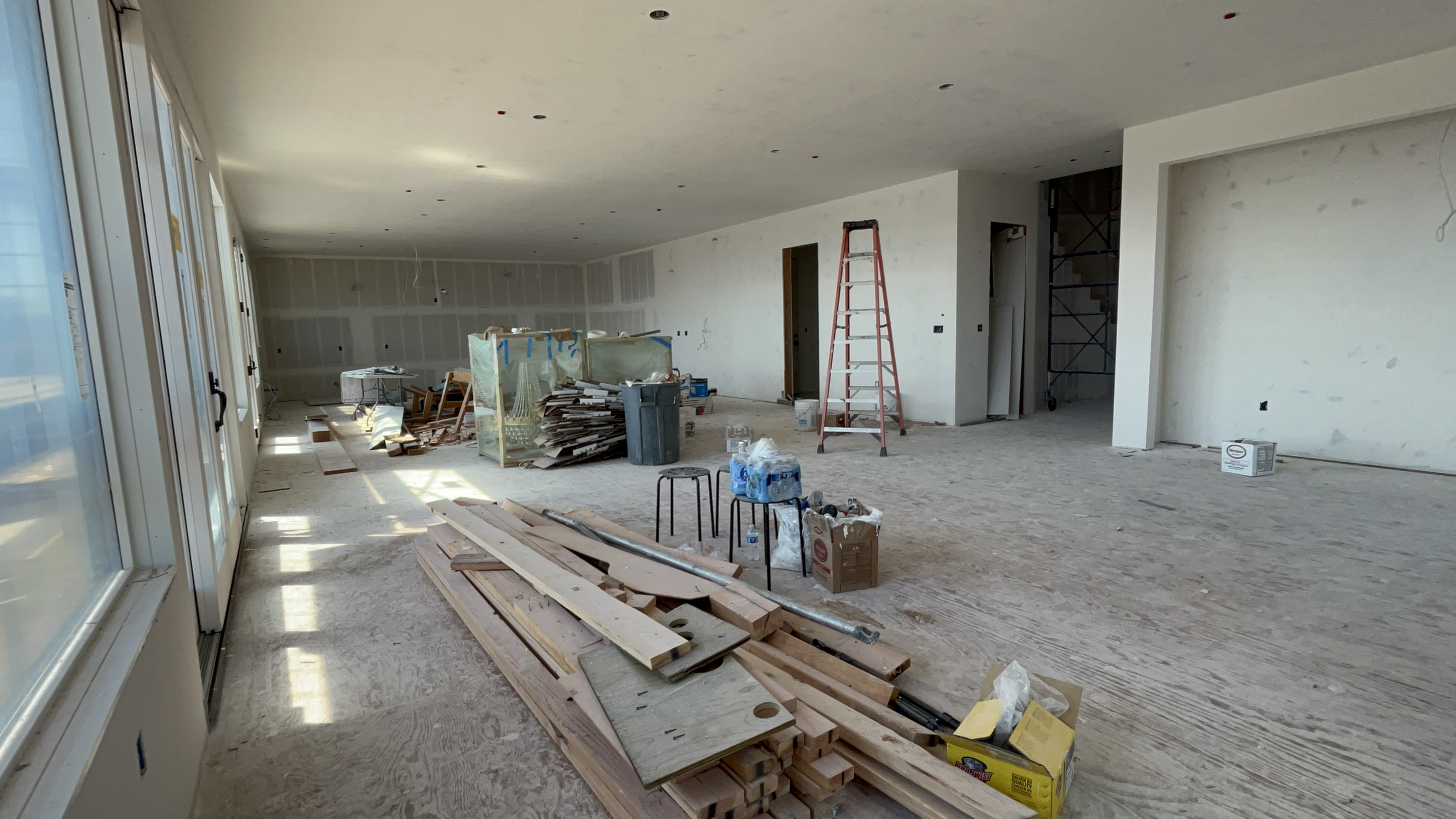 This image shows a spacious, unfinished interior room under construction, likely a living area or open-concept living space. The room features bare drywall, plywood subflooring, and exposed construction materials, including stacked lumber and a ladder, indicating an active renovation project. Large windows on the left allow for ample natural light, highlighting the potential for a bright and open floor plan once the finish work is completed.