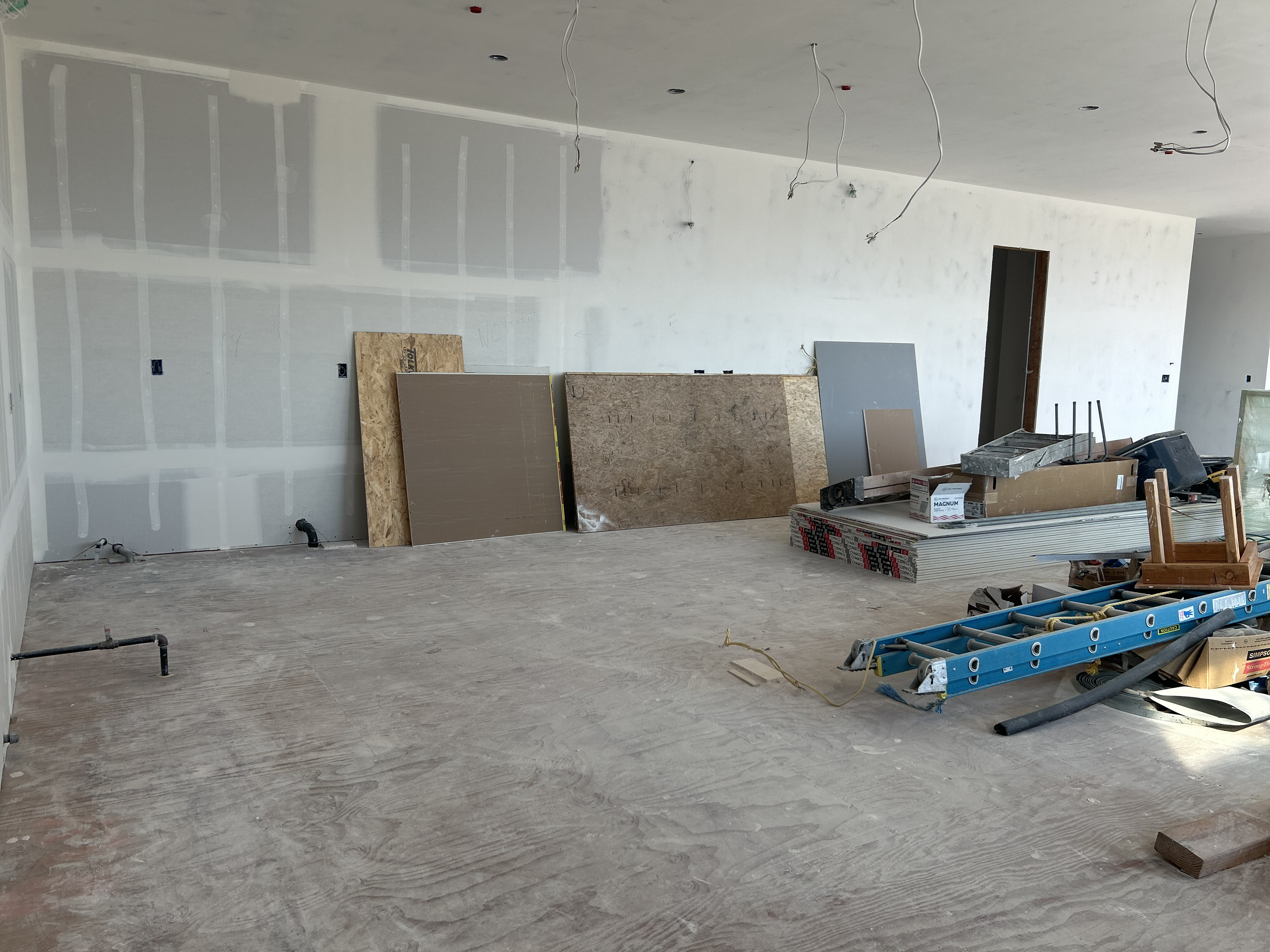 This image depicts an unfinished interior space, likely a kitchen under renovation, evidenced by the exposed plumbing and electrical wiring. Construction materials, including drywall sheets and plywood, are stacked against a wall alongside a ladder and various tools. The area features bare plywood flooring and unfinished drywall, presenting a blank canvas for customization and highlighting the potential for a modern renovation project.