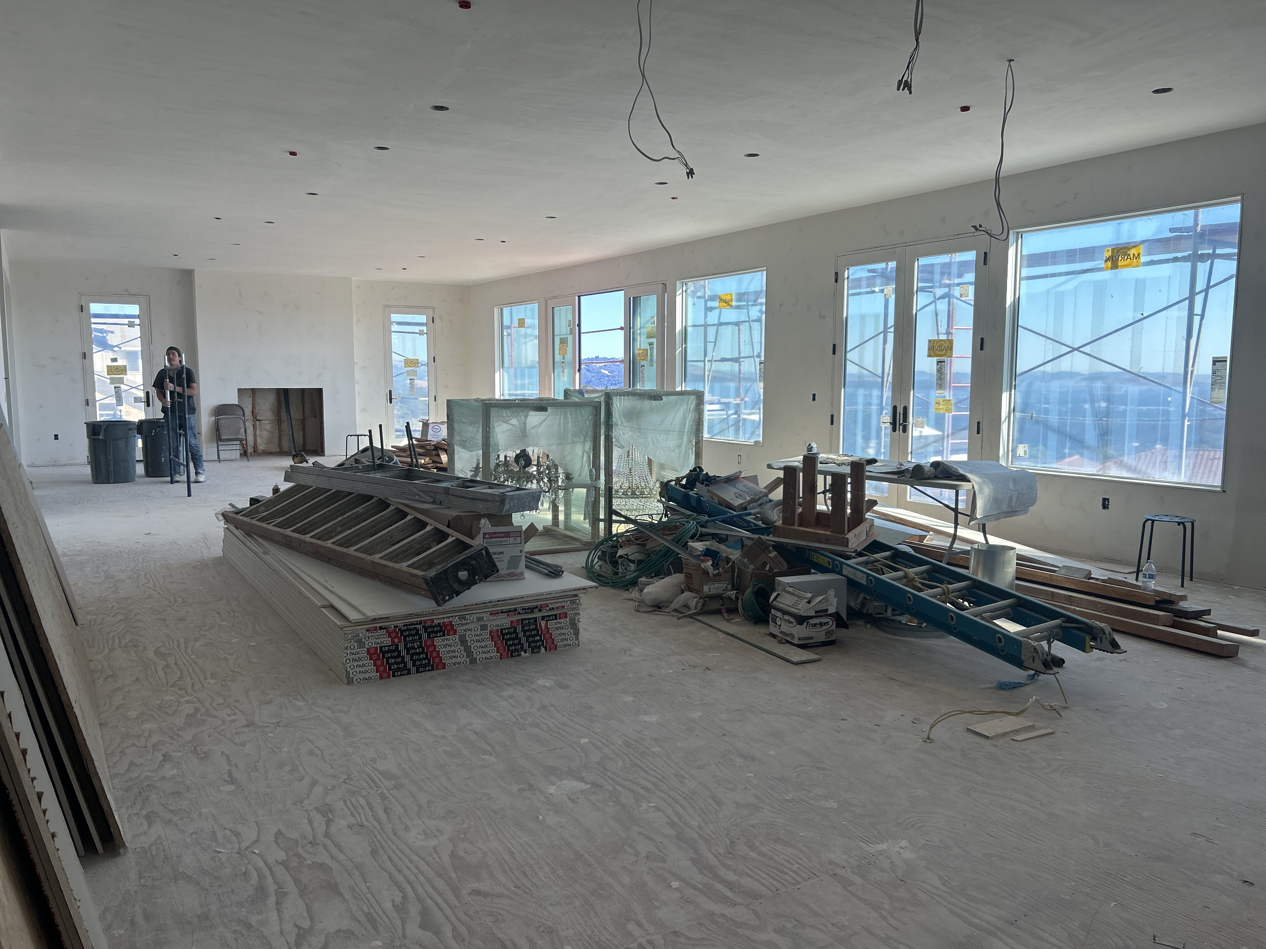 This is a large, unfinished interior space under construction, likely intended as a living room or great room. It features expansive windows with panoramic views, smooth-finished white walls, and a plywood subfloor, reflecting a high-end property in the middle of being built. The space is currently being used for storage of building materials, including ladders, drywall, and a crystal chandelier in a protective crate.