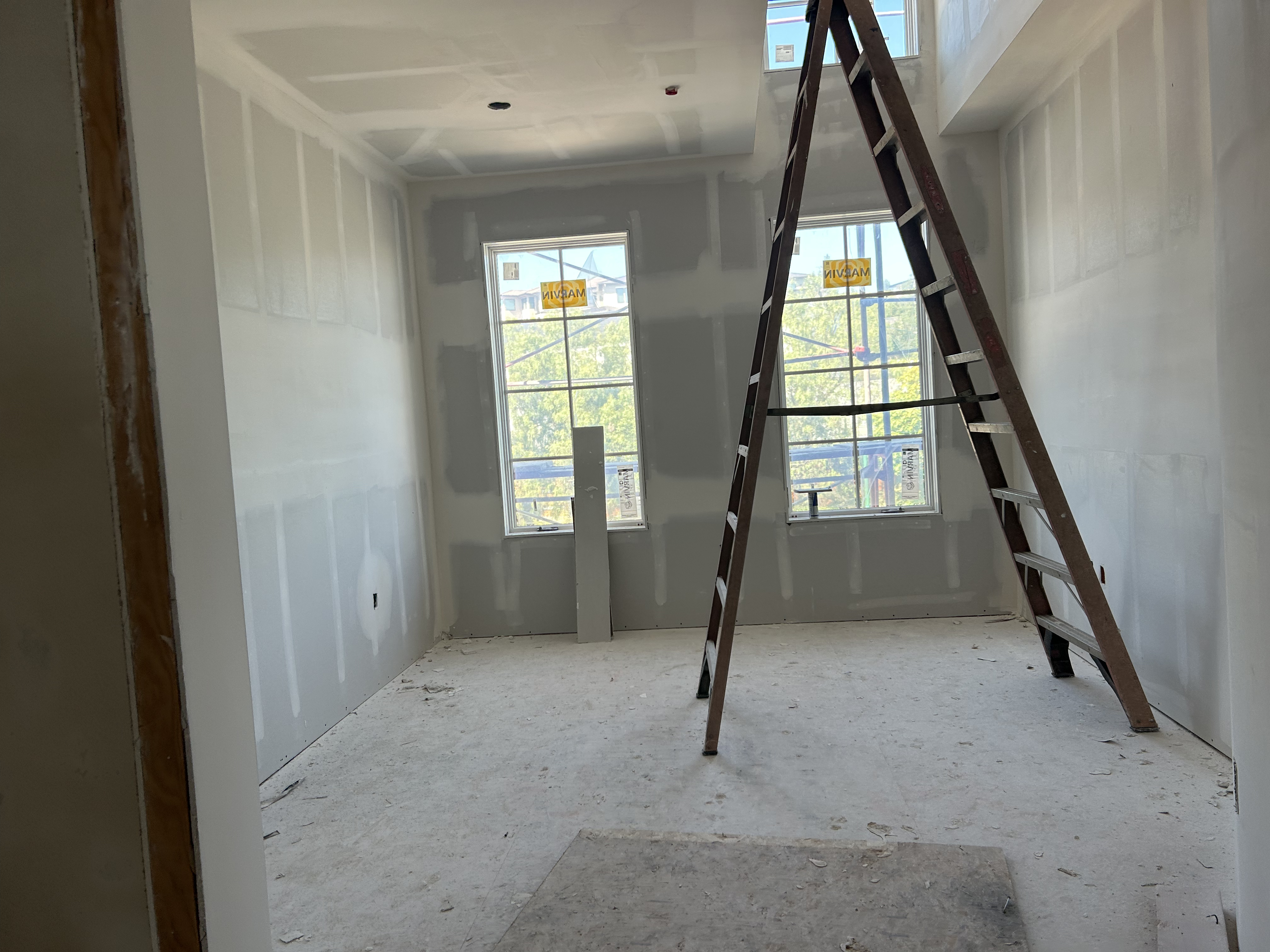 This image showcases a room under construction, likely intended as a bedroom or office, featuring unfinished drywall with visible mudding on the seams. A prominent wooden A-frame ladder dominates the center of the space, which includes two windows with outdoor views and a ceiling skylight. The room is in an early stage of finish, providing a clean slate for future flooring, paint, and trim installation.