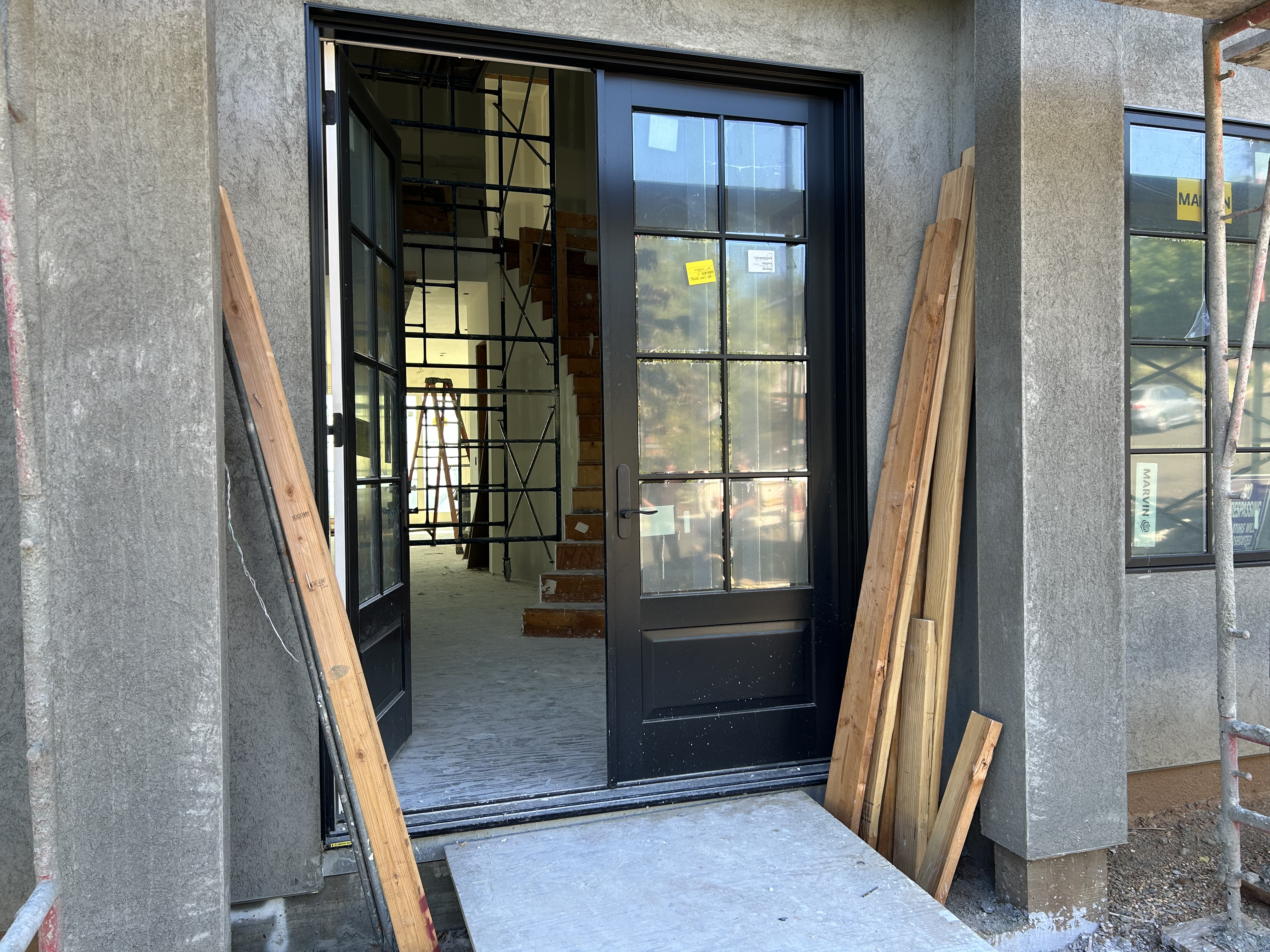 This image shows the entryway of a house currently under construction, featuring a sleek, black-framed glass double door that opens into an unfinished interior space. The exterior is covered in textured gray stucco, and wooden scaffolding and lumber are positioned to the side, indicating an active building project. The entrance provides a glimpse into the home's framing and staircase, highlighting the potential for a modern, high-end design.