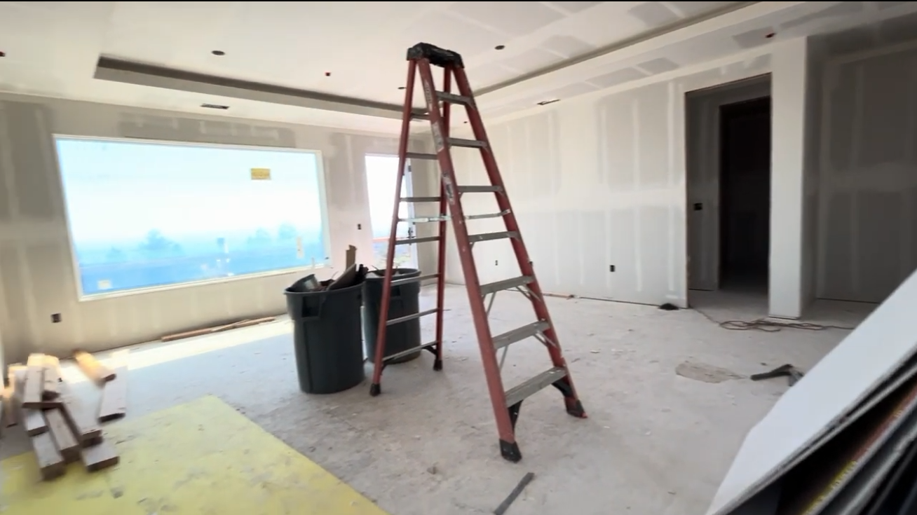 This interior image shows a spacious, unfinished living room currently under construction. Key features include freshly installed drywall that has been taped and mudded, a large window letting in natural light, and a high ceiling with recessed lighting. The room is currently occupied by a large A-frame stepladder and construction debris, providing a blank canvas for potential buyers to envision their own design.