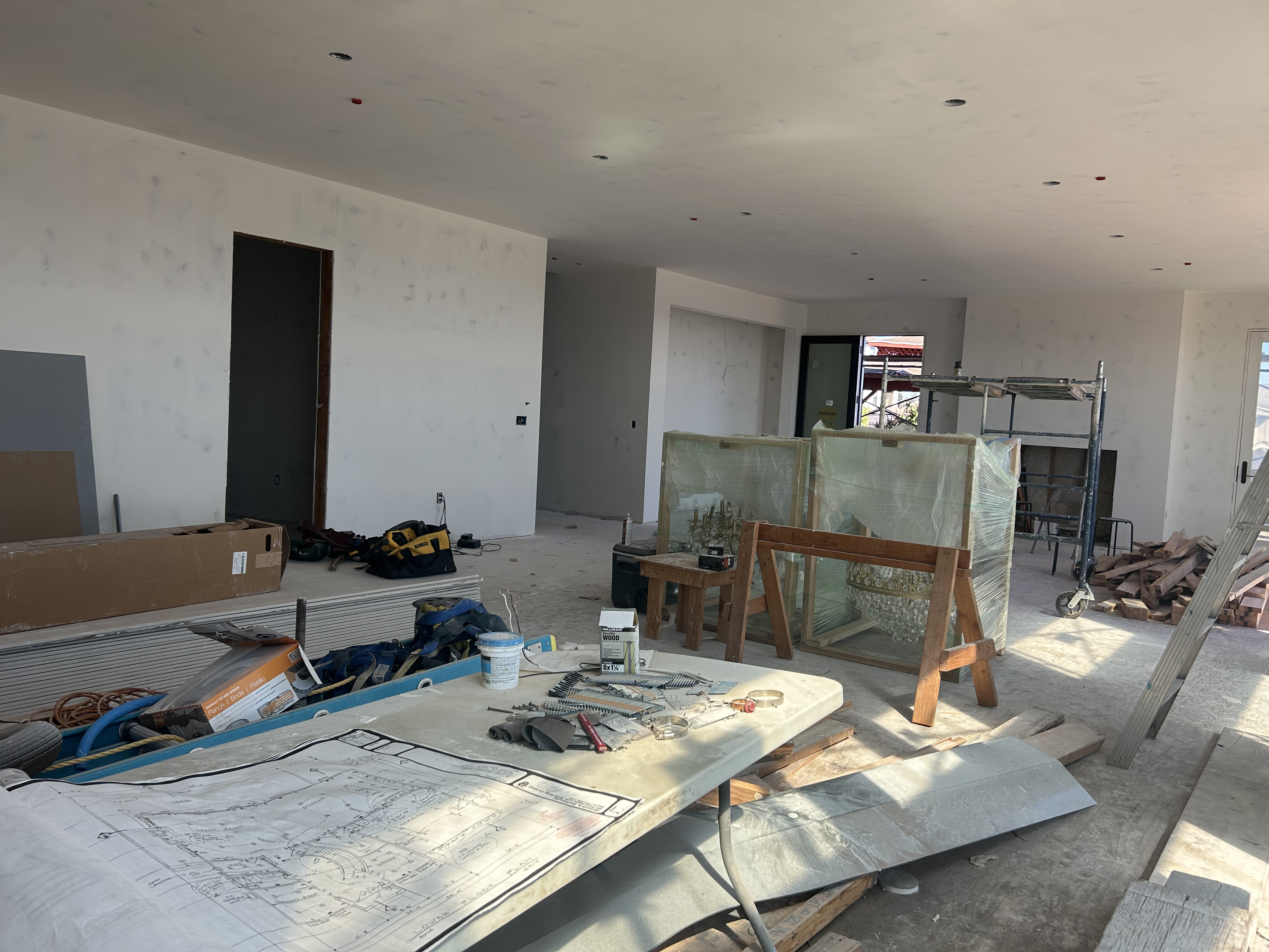 This image shows the interior of a house currently under construction in a state of renovation or build-out. The space features unfinished drywall, exposed wall studs, and various construction supplies, materials, and equipment such as scaffolding, tools, and stacked lumber. While the room layout is open and spacious with modern entryways and high ceilings with recessed lighting, the presence of plastic-wrapped items and blueprints on a temporary table highlights that the home is not yet move-in ready.
