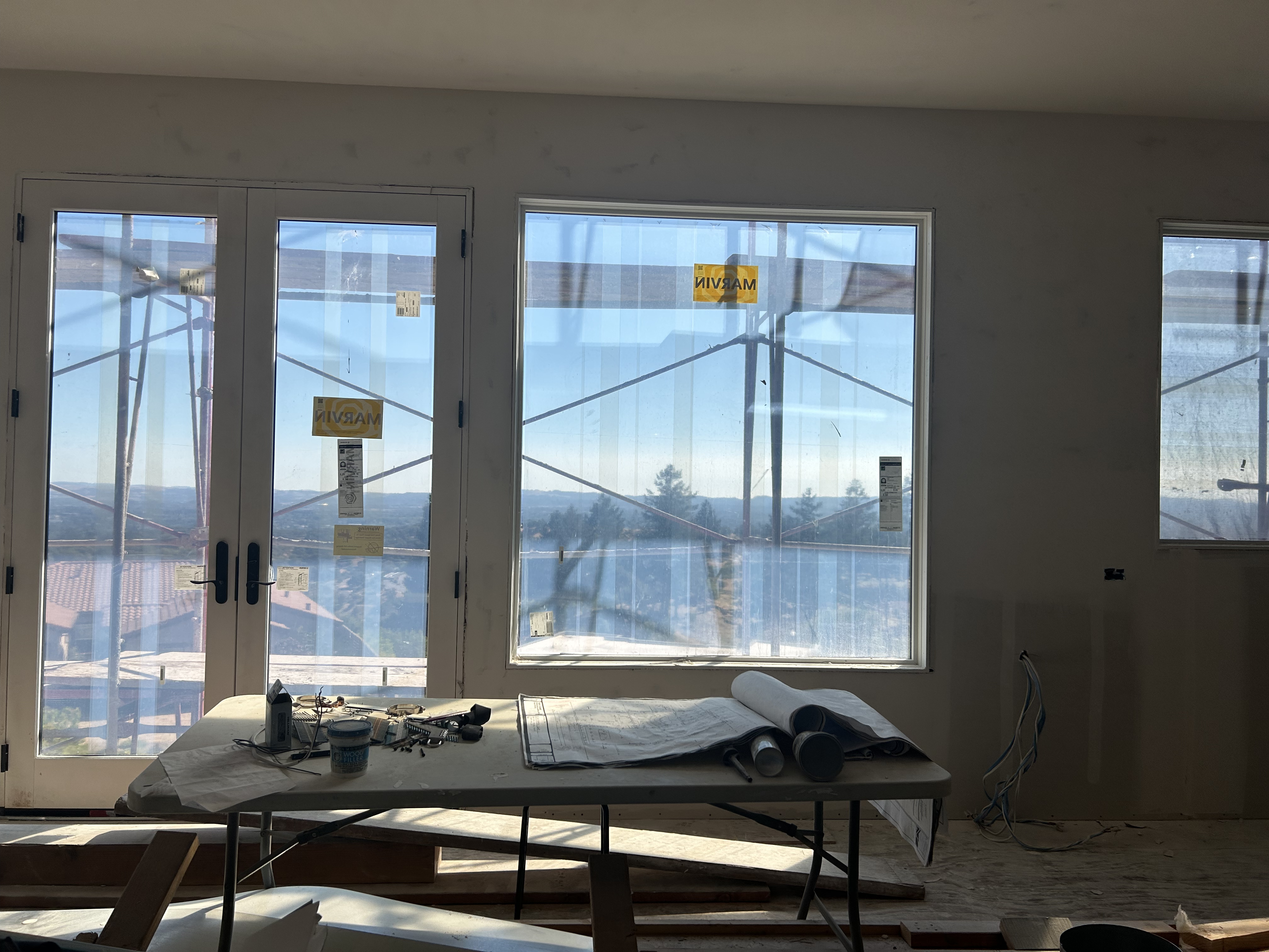 This interior space is currently under construction, featuring bare walls and large, energy-efficient windows that offer expansive views of the surrounding landscape and scaffolding. A folding table sits in the center holding construction plans and tools, highlighting the ongoing work in what appears to be a living or gathering area. The open floor plan and significant glazing promise a bright, airy environment with potential for impressive indoor-outdoor connectivity.
