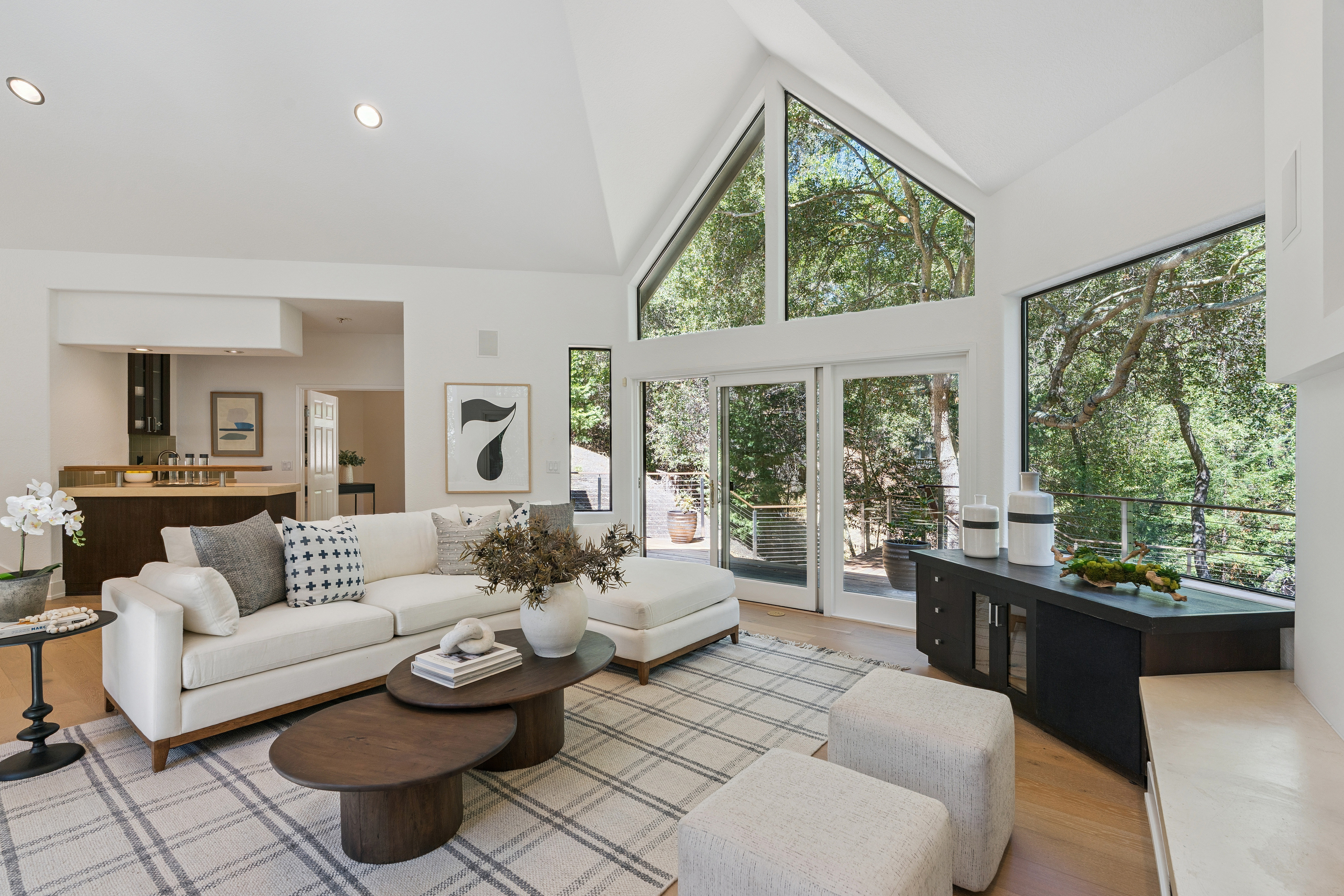 This bright and airy living room features a vaulted ceiling and expansive windows that provide a scenic view of the surrounding trees. The space is furnished with a comfortable white sectional sofa, a pair of dark wood nesting coffee tables, and two neutral-toned ottomans atop a plaid area rug. A dark sideboard sits against the wall, and the open-concept layout flows seamlessly into a kitchen area in the background.