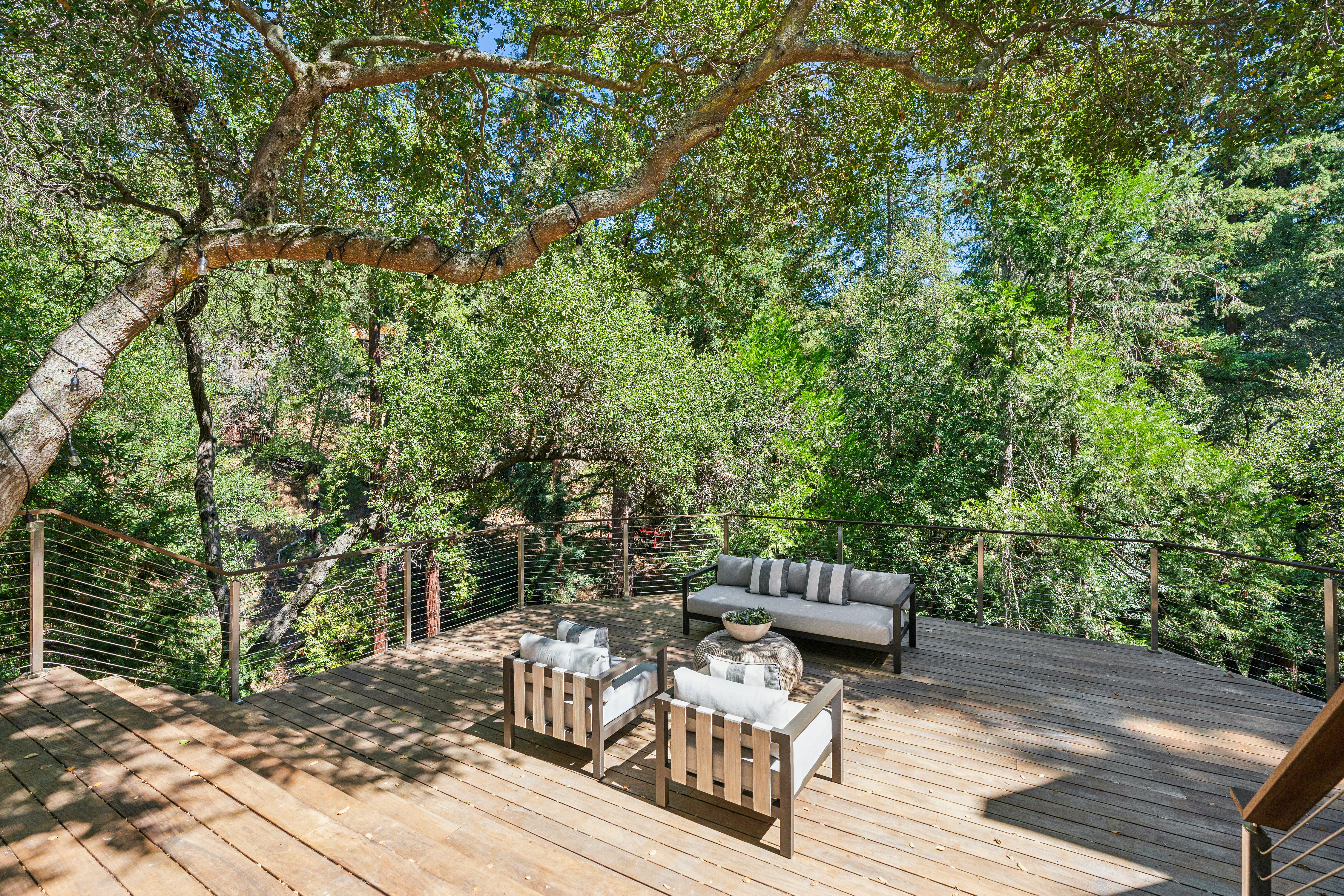 This elevated wooden deck offers a serene outdoor living space nestled among mature trees, providing a private, forest-like atmosphere. The area is furnished with a modern sofa and two matching armchairs arranged around a circular coffee table, all set against a backdrop of lush greenery. A sleek cable railing system ensures unobstructed views of the surrounding woodland, creating a peaceful and inviting retreat.