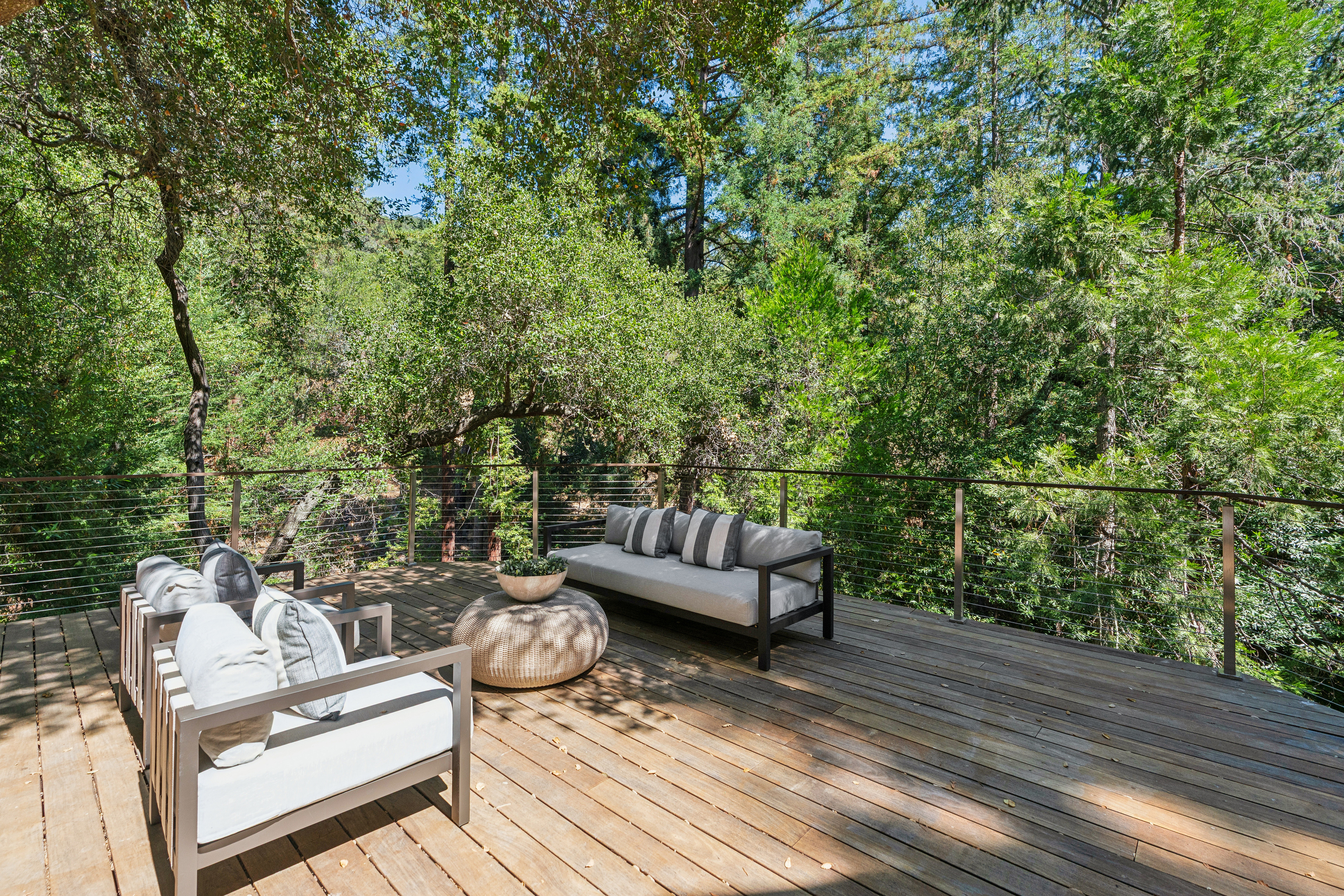 This elevated wooden deck offers a serene outdoor living space nestled among lush, mature trees. The area is furnished with a modern sofa and matching armchairs, centered around a round woven coffee table, creating an inviting atmosphere for relaxation. A sleek metal cable railing provides safety while maintaining an unobstructed view of the surrounding forest canopy.