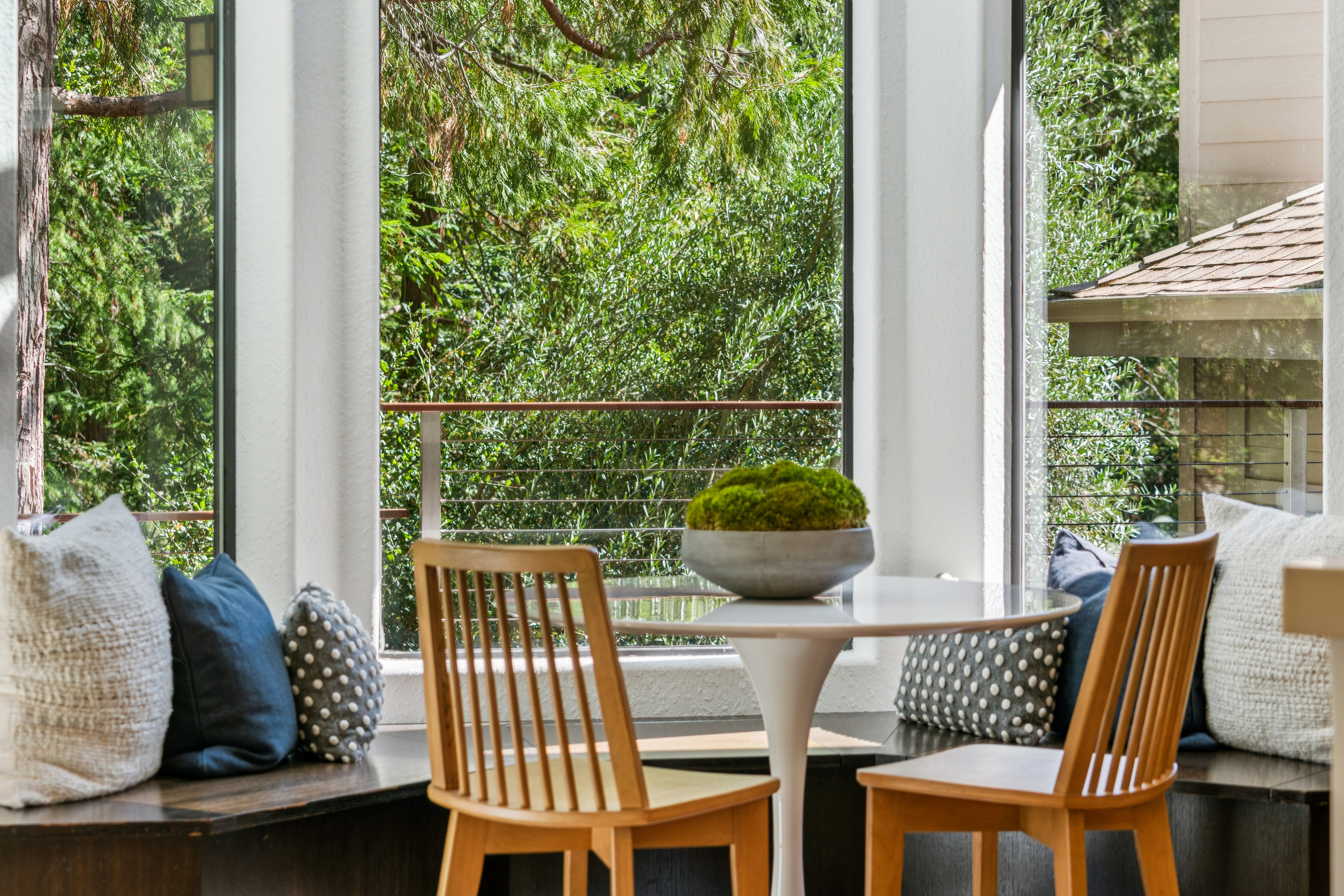 This bright dining nook features a circular white pedestal table centered in front of large windows that offer a serene view of lush greenery and a balcony. Built-in bench seating lines the window wall, accented with a mix of solid blue and polka-dot throw pillows, while two light wood chairs provide additional seating. A decorative bowl filled with moss sits as a centerpiece, creating a peaceful and inviting atmosphere perfect for casual meals.