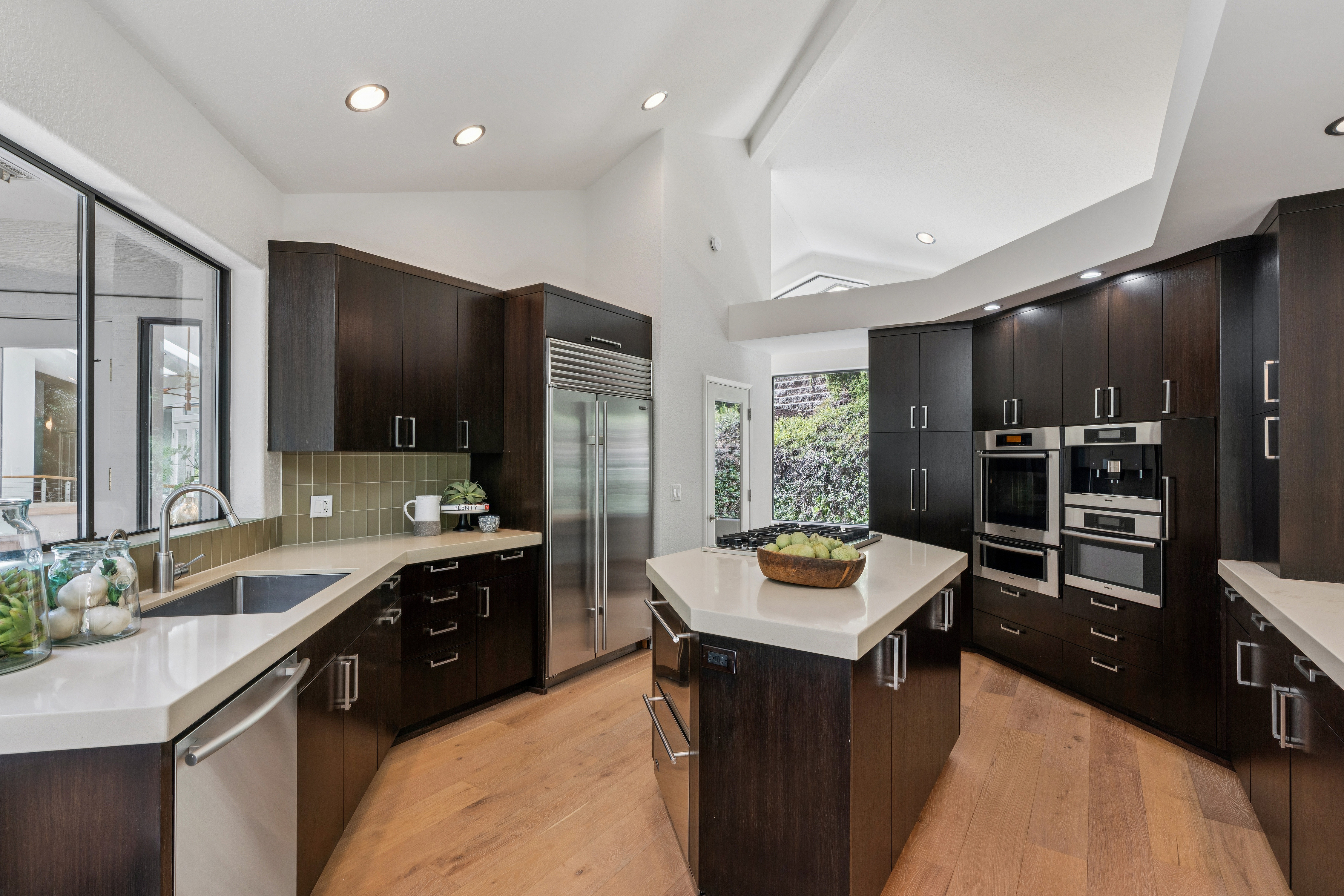 This modern kitchen features dark wood cabinetry contrasted by light-toned stone countertops and a central island. The space is equipped with high-end stainless steel appliances, including a built-in refrigerator and a wall-mounted oven and coffee system. Large windows and a vaulted ceiling create an airy, open atmosphere, while the light wood flooring adds warmth to the sophisticated design.