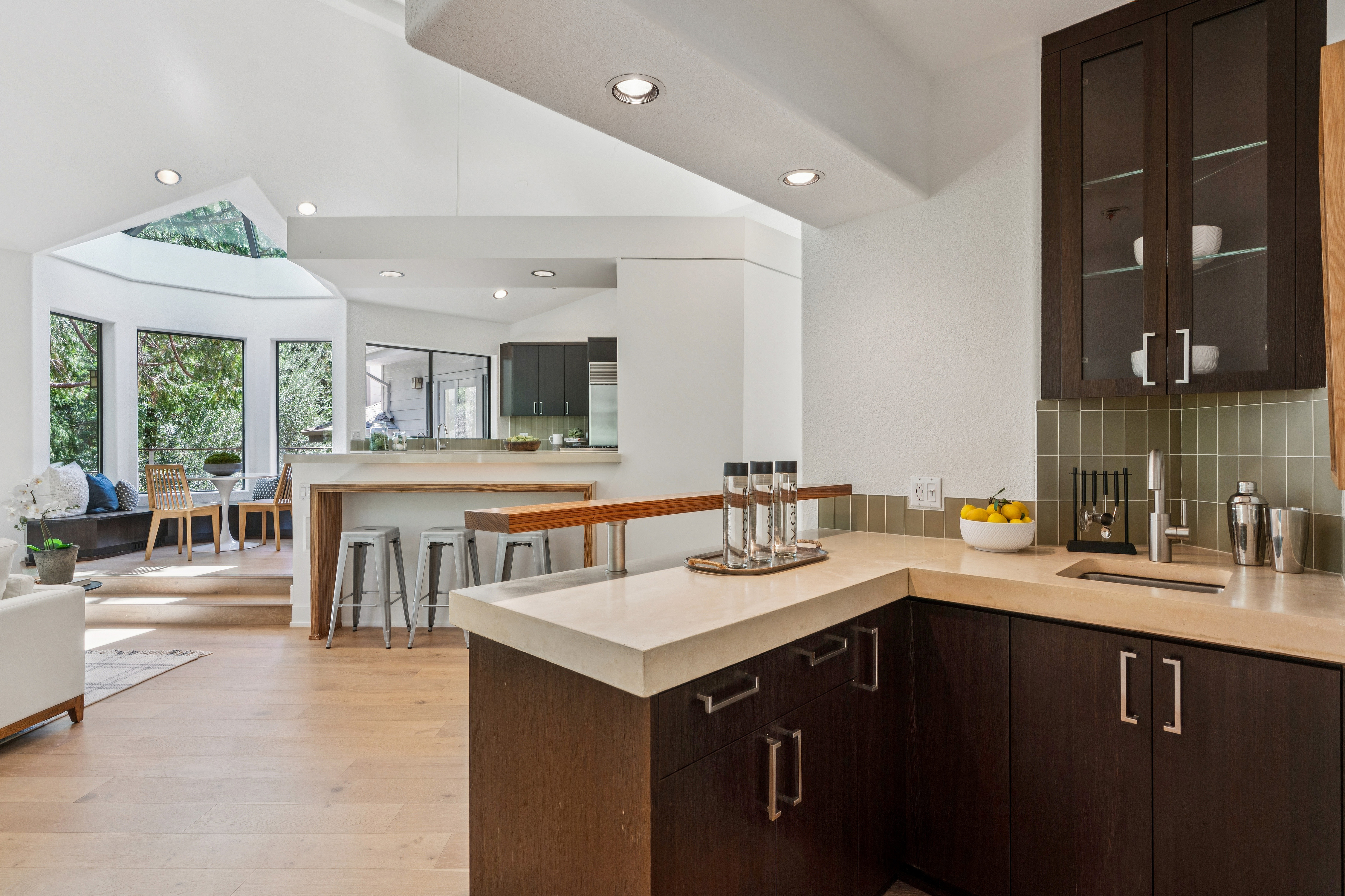 This bright, open-concept kitchen features dark wood cabinetry contrasted by light-toned stone countertops and a tiled backsplash. The space flows seamlessly into a sun-drenched breakfast nook with large windows and a skylight, creating an airy and inviting atmosphere. A raised bar area with stools provides additional seating, while recessed lighting highlights the clean lines and modern aesthetic of the home.