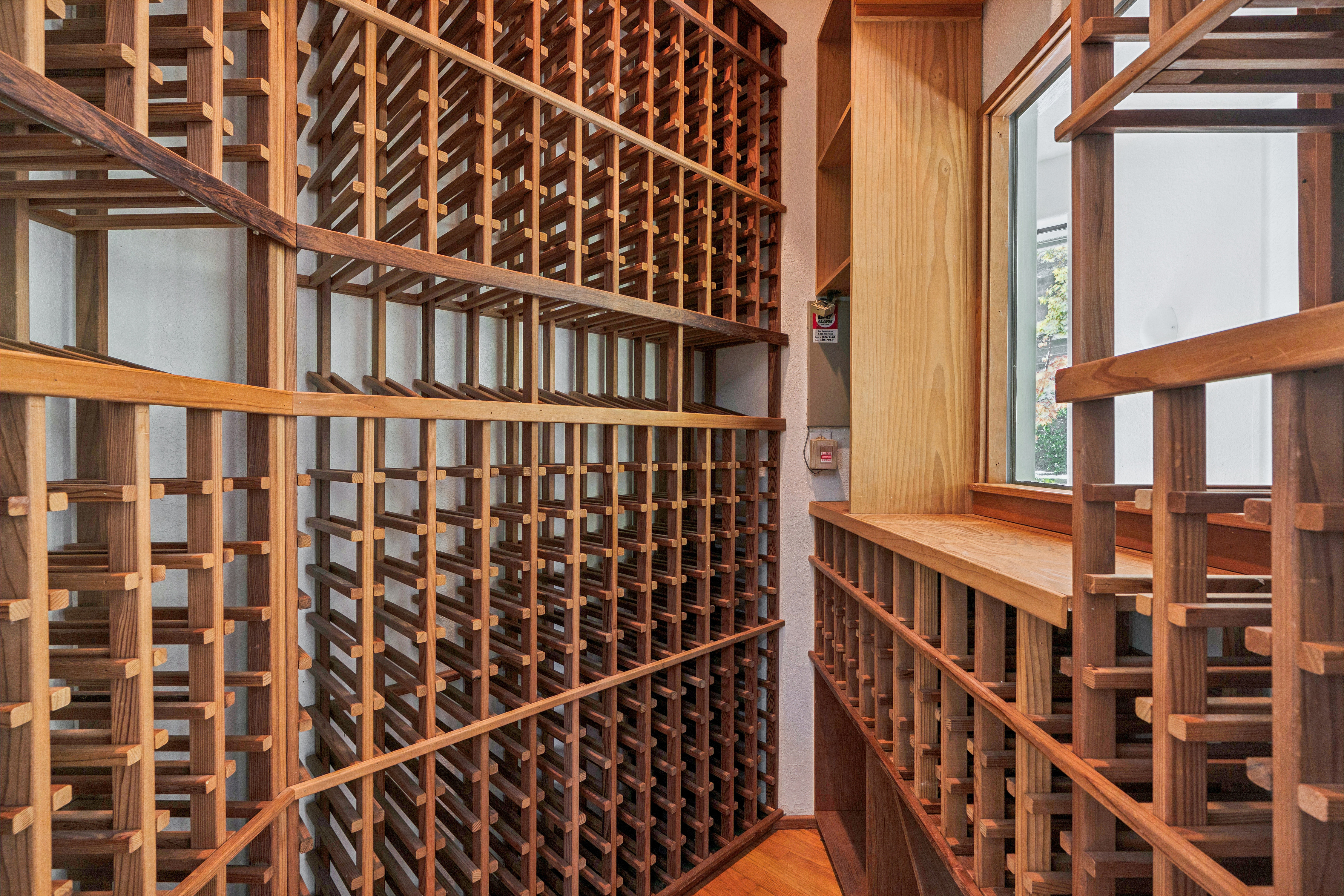 This dedicated wine cellar features extensive custom-built wooden racking designed for organized bottle storage. The space includes a built-in counter area and a window that allows natural light to filter into the room, creating a functional and sophisticated environment for wine enthusiasts. The warm wood tones and structured layout provide a classic, professional aesthetic suitable for a high-end home.
