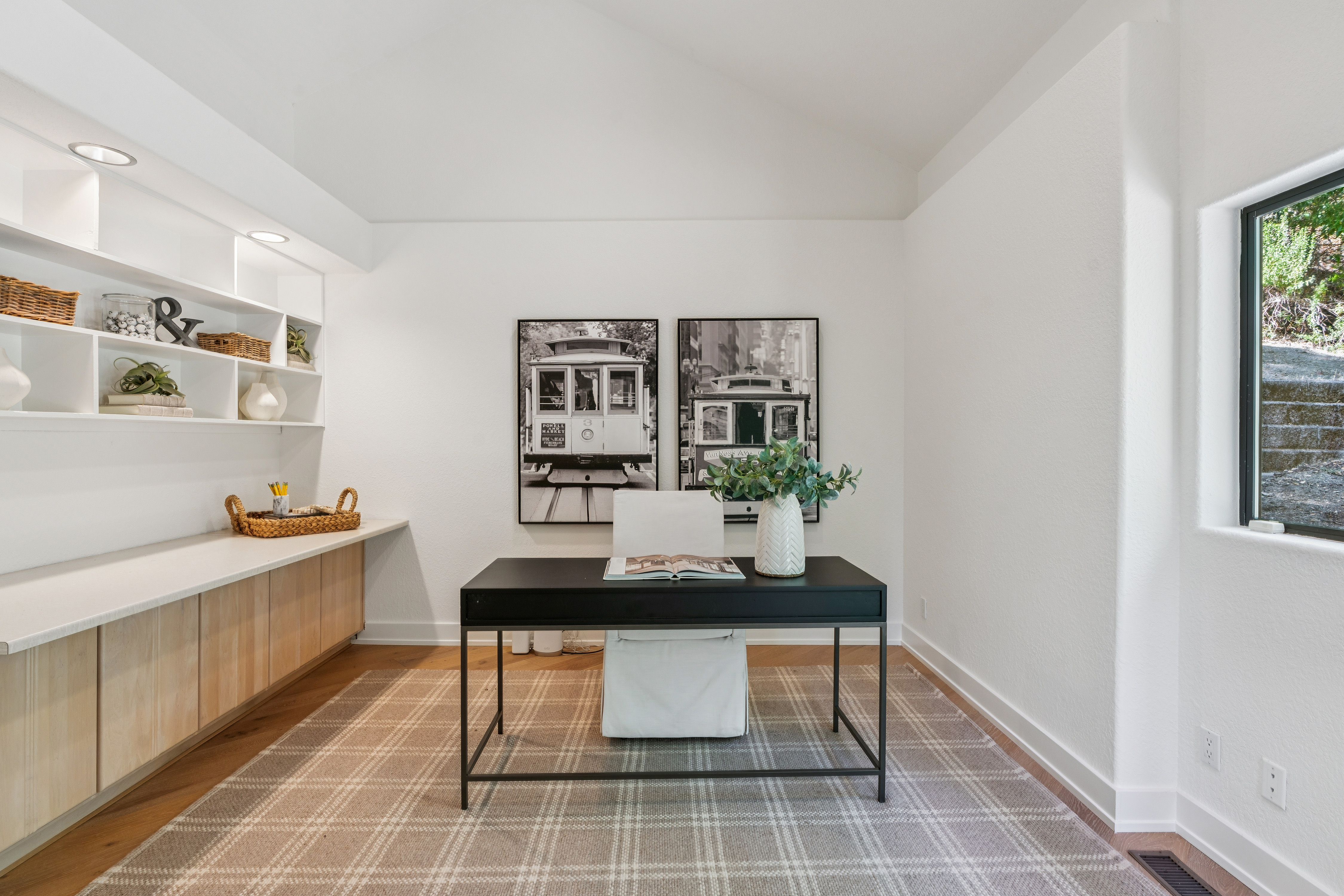 This bright and modern home office features a minimalist black desk centered in the room, paired with a white upholstered chair. The space is accented by built-in light wood cabinetry with open shelving and two large black-and-white framed prints of cable cars on the wall. A neutral plaid area rug anchors the room, while a window on the right provides natural light and a view of the exterior landscape.