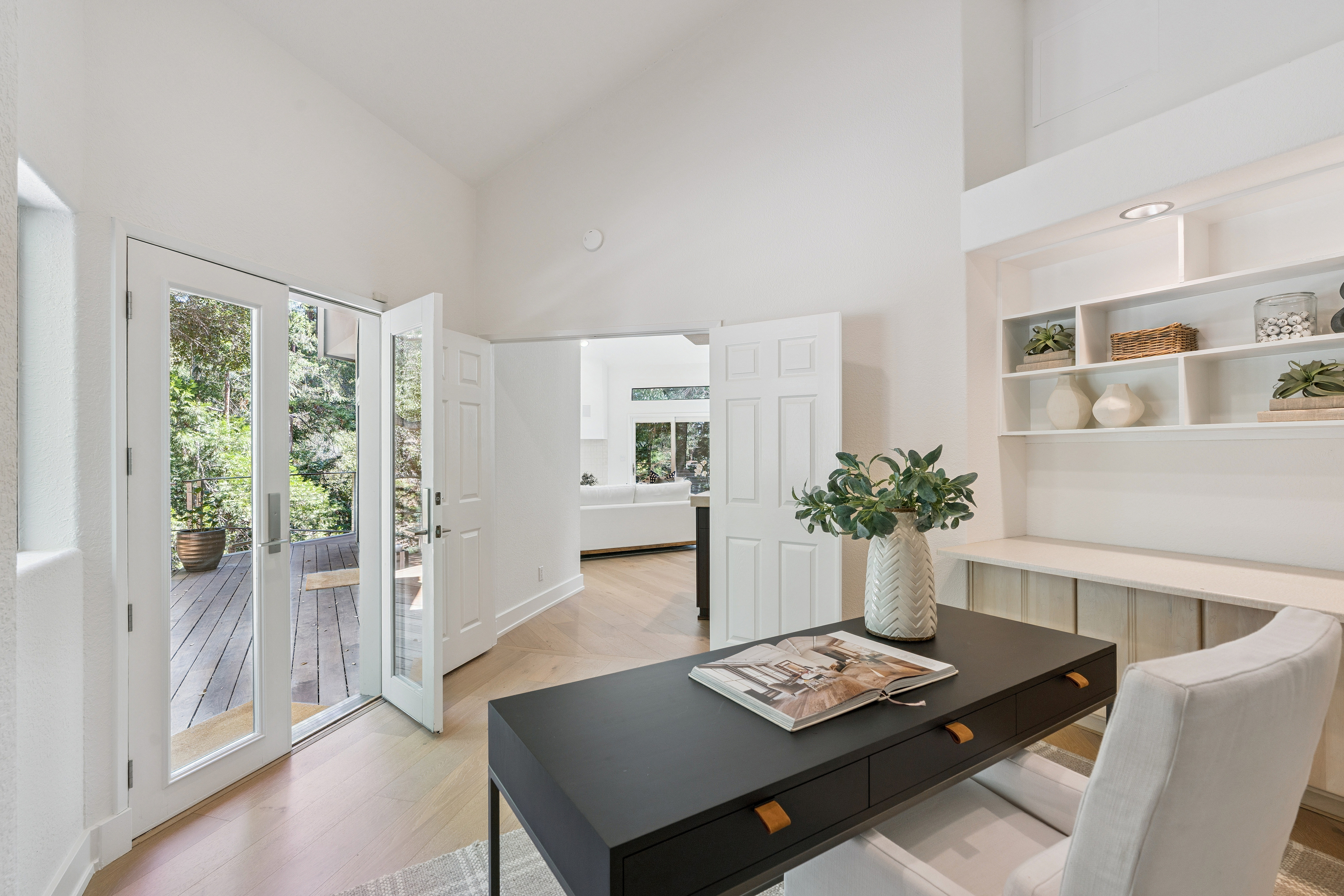 This bright home office features a sleek black desk centered in the room, complemented by a white upholstered chair and built-in shelving units. Double glass doors open directly onto a wooden deck, seamlessly blending the indoor workspace with the lush greenery outside. The room is finished with light wood flooring and crisp white walls, creating a clean, professional, and inviting atmosphere.