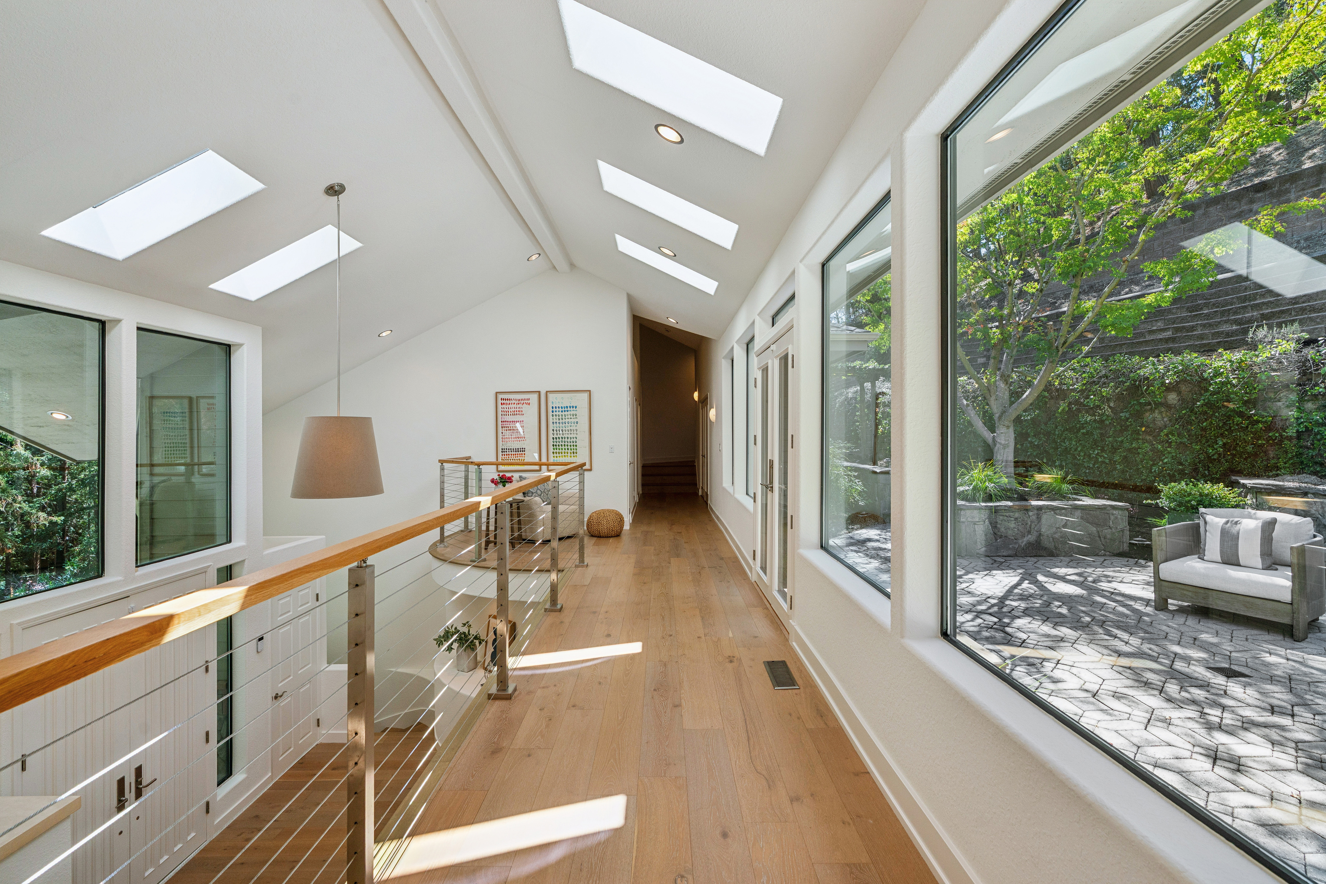 This bright, airy hallway features light wood flooring and a vaulted ceiling punctuated by multiple skylights that flood the space with natural light. A sleek railing with horizontal cables borders the staircase, while large windows along the right wall provide a seamless connection to the outdoor patio and garden. The perspective captures the length of the corridor, emphasizing the open, modern architectural design and the transition between indoor and outdoor living spaces.