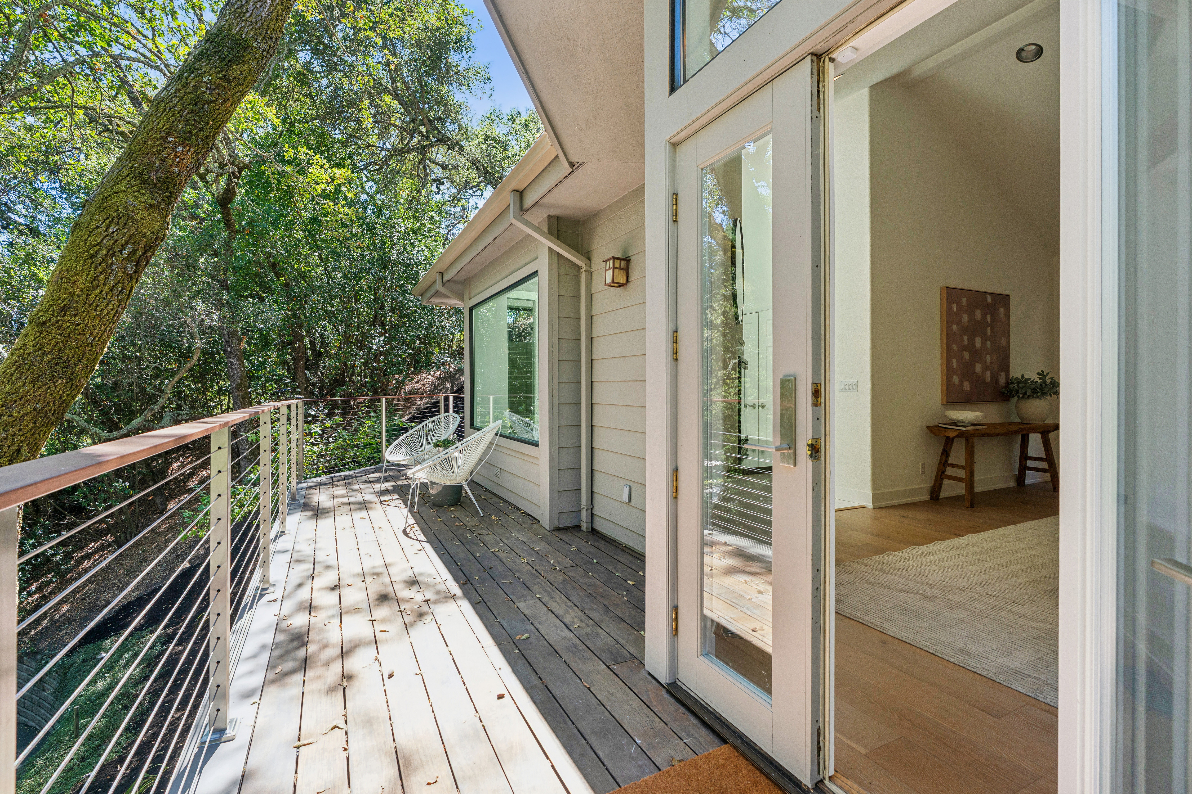 This image captures a serene wooden deck overlooking a lush, tree-filled landscape, accessible through a glass-paned door. The deck features modern wire-railing and two white, woven-style chairs, creating a peaceful outdoor seating area. The perspective looks out from the threshold of the home, highlighting the seamless transition between the interior living space and the natural surroundings.