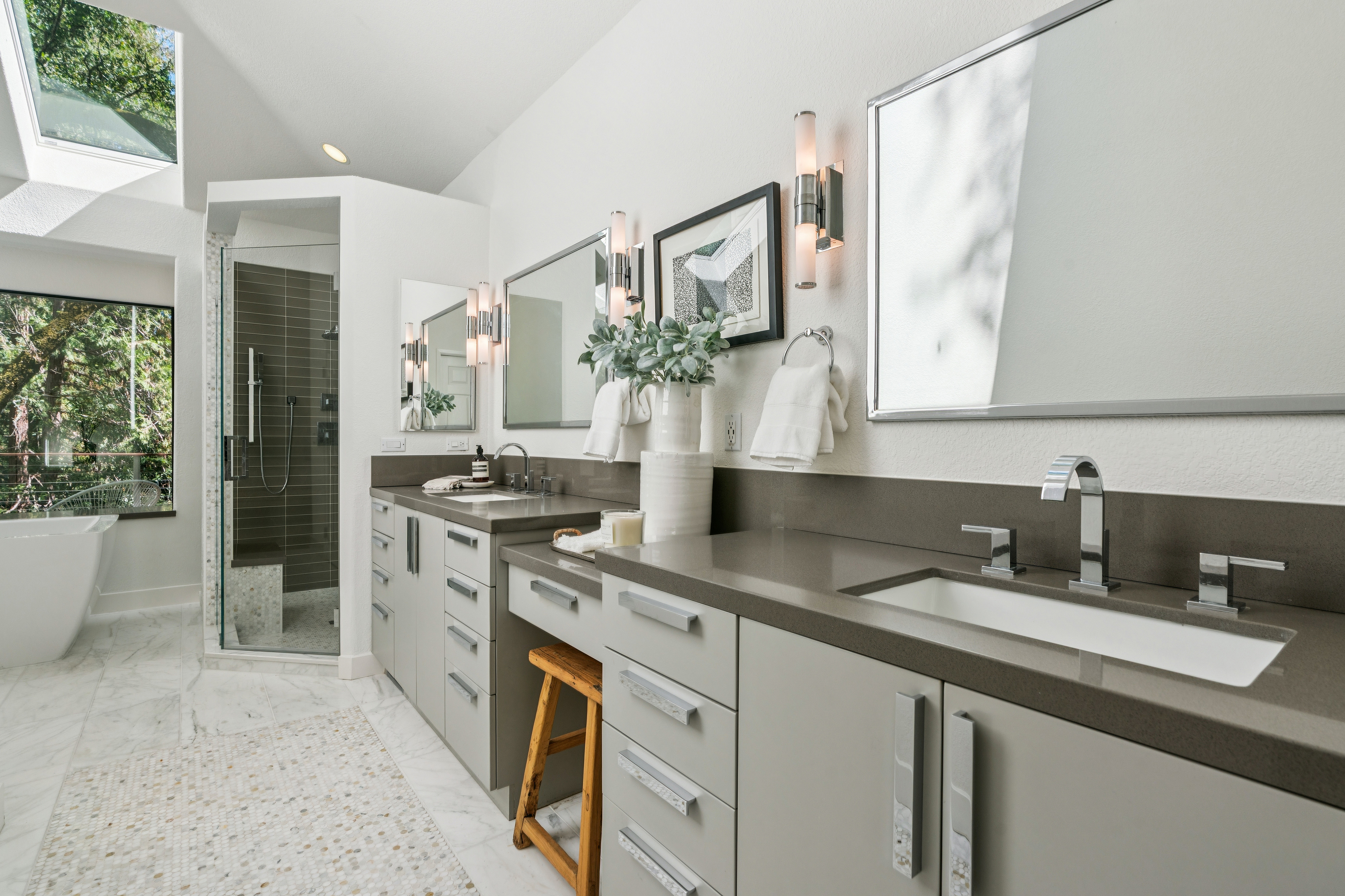 This primary bathroom features a dual-vanity setup with light-toned cabinetry, dark stone countertops, and modern chrome fixtures. A glass-enclosed shower with dark tile walls sits to the left, while a freestanding tub is visible near a large window that provides natural light. The space is finished with light-colored flooring and a decorative rug, creating a bright and contemporary atmosphere.