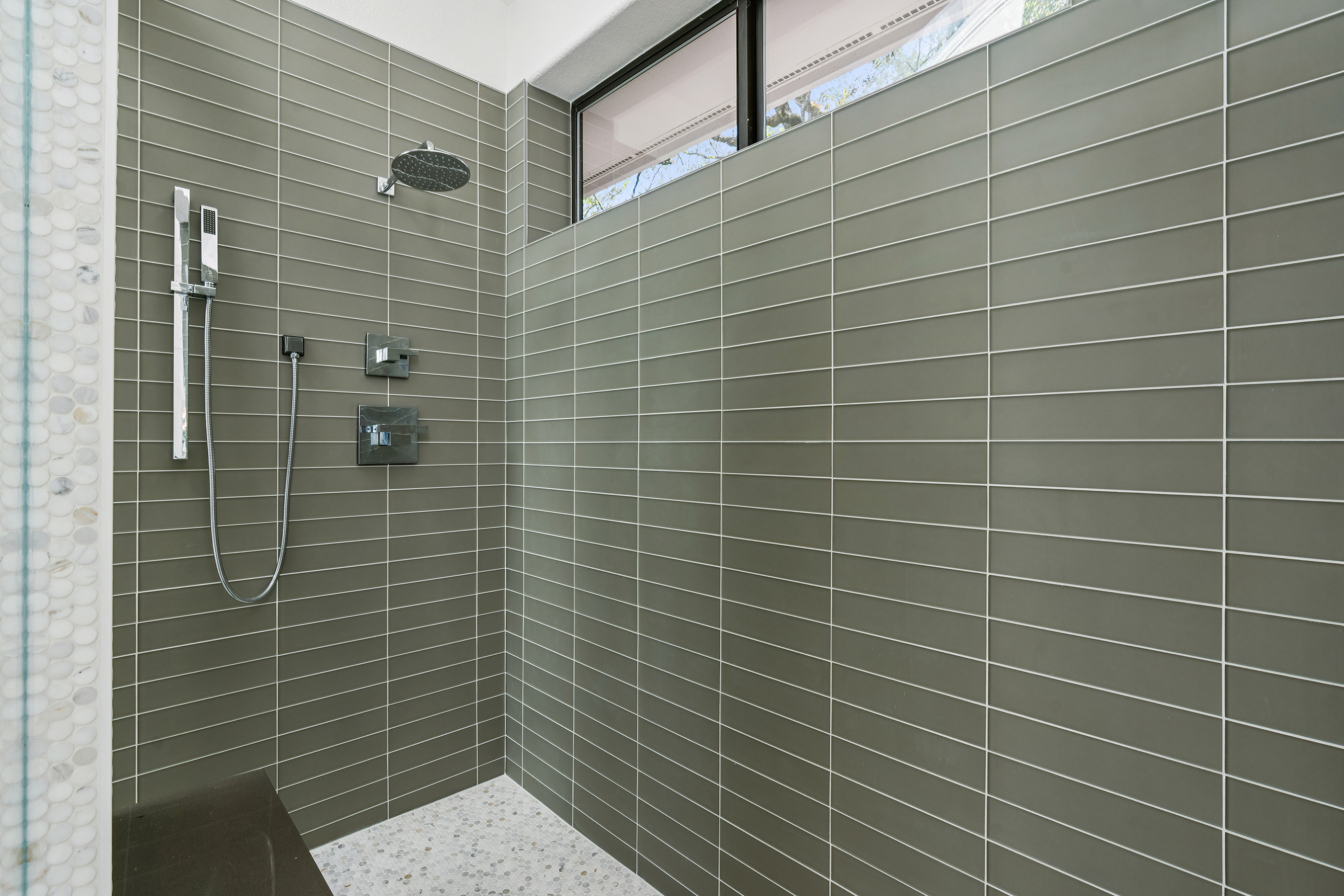 This modern shower space features long, rectangular olive-green wall tiles arranged in a horizontal pattern, creating a sleek and contemporary aesthetic. A rainfall showerhead and a separate handheld sprayer are mounted on the wall, complemented by a small, high-placed window that allows natural light to filter in. The shower floor is finished with small, circular mosaic tiles, and a dark bench is visible in the corner, adding a functional and sophisticated touch to the design.
