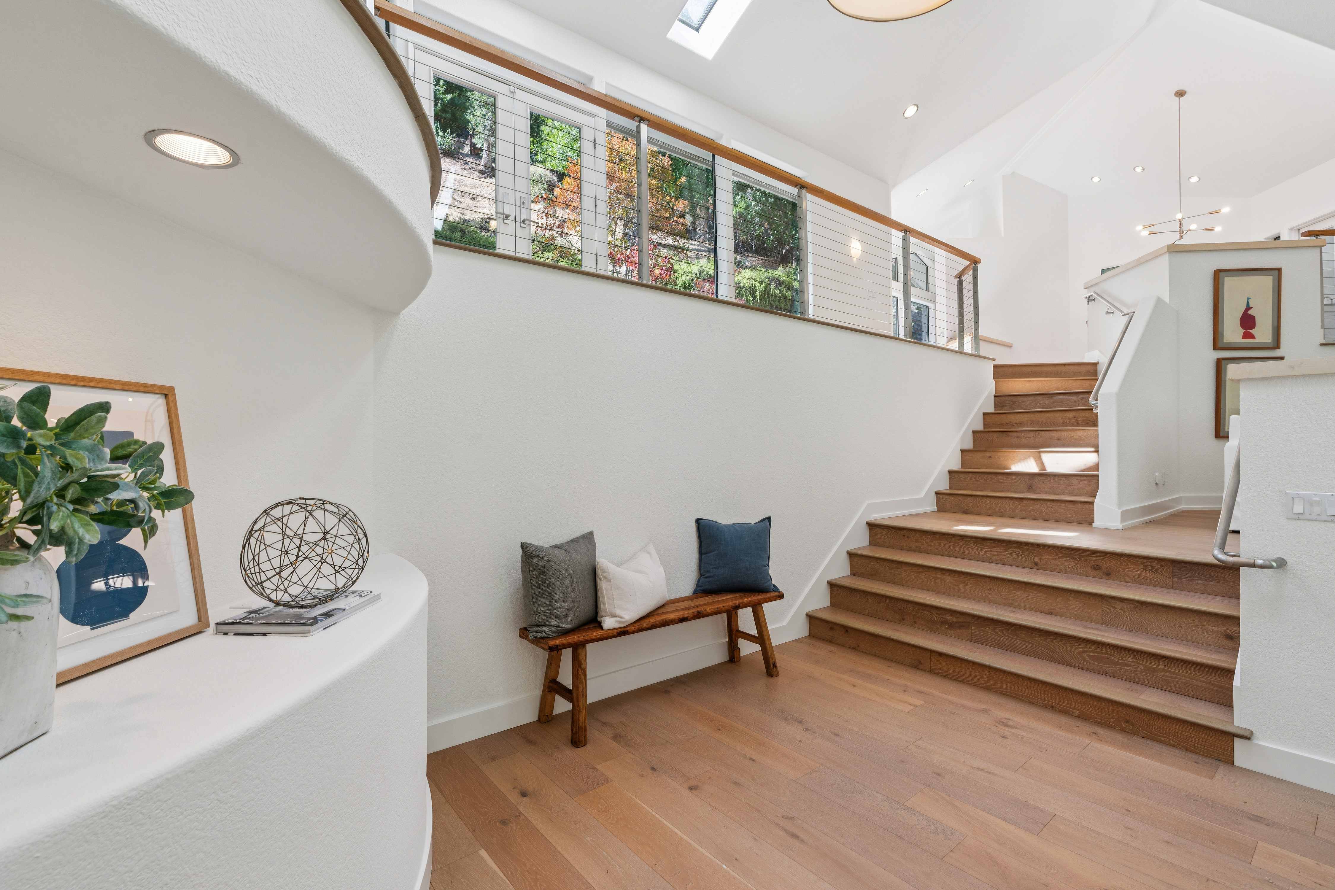 This bright and airy entryway features a staircase with light wood treads leading to an upper level, complemented by a sleek metal railing. A rustic wooden bench with decorative pillows sits against a crisp white wall, while a curved architectural ledge displays a potted plant and a geometric sculpture. The space is illuminated by natural light streaming through a series of windows above the landing and recessed ceiling lighting, creating a welcoming and modern atmosphere.