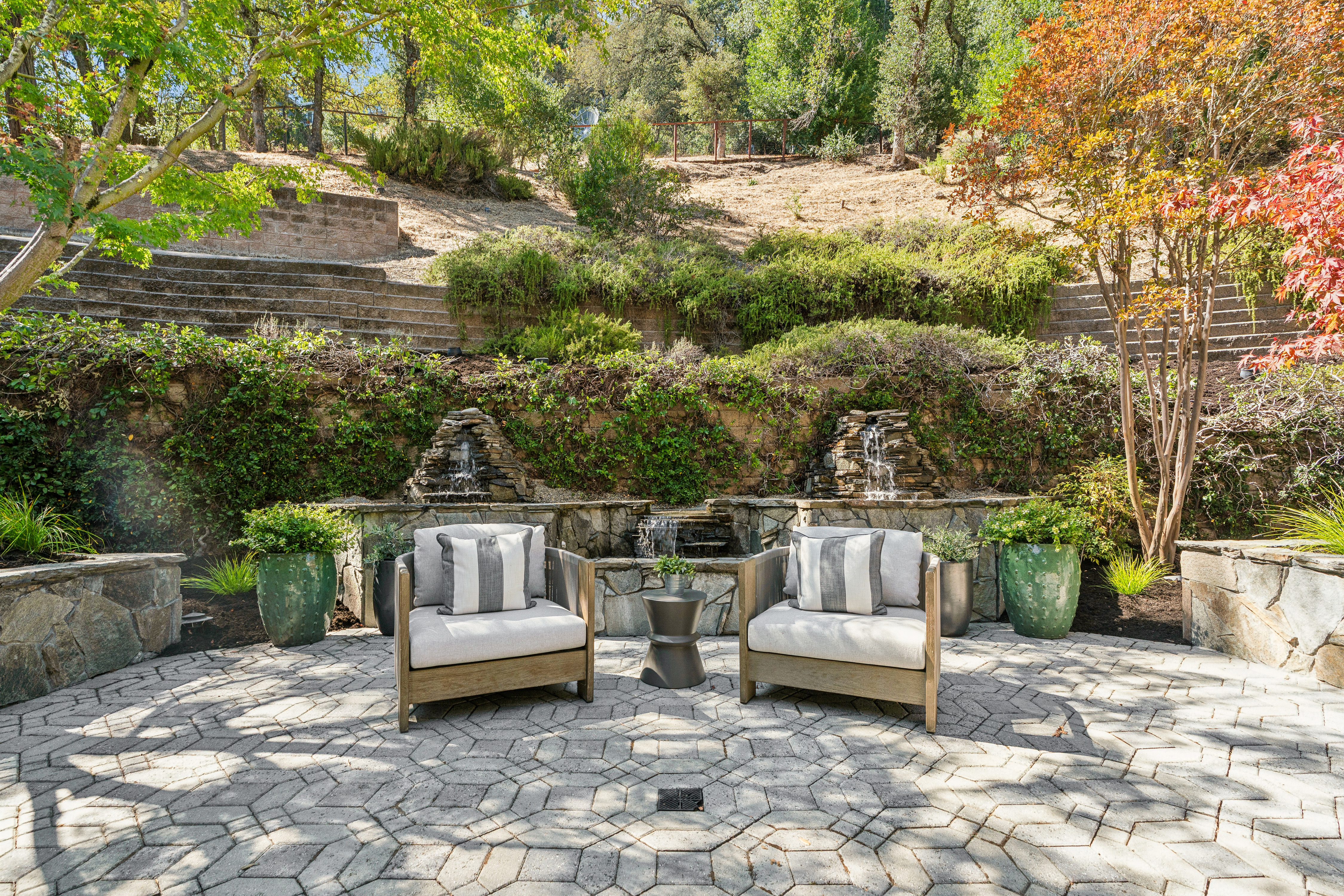 This serene outdoor patio features a geometric stone paver floor and two comfortable armchairs centered around a small side table. The space is anchored by a tiered stone water feature built into a retaining wall, surrounded by lush greenery and terraced landscaping. The setting offers a peaceful, private retreat with a sophisticated, natural aesthetic.