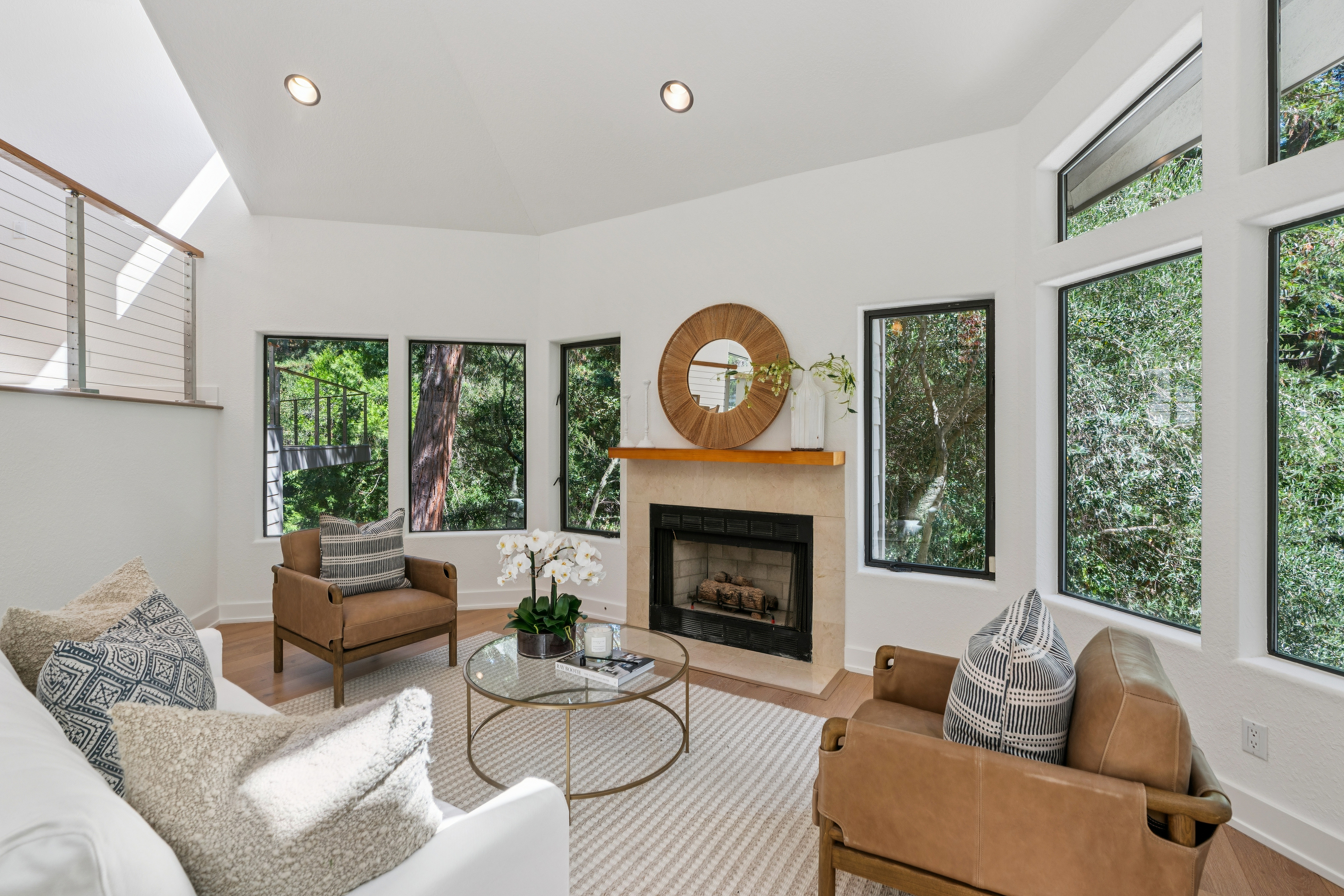This bright and airy living room features a vaulted ceiling and an abundance of large windows that frame lush, green outdoor views. The space is anchored by a central fireplace with a stone surround and a wood mantel, complemented by two leather armchairs and a white sofa arranged around a glass-topped coffee table. The neutral color palette and natural light create a serene, inviting atmosphere perfect for relaxation.
