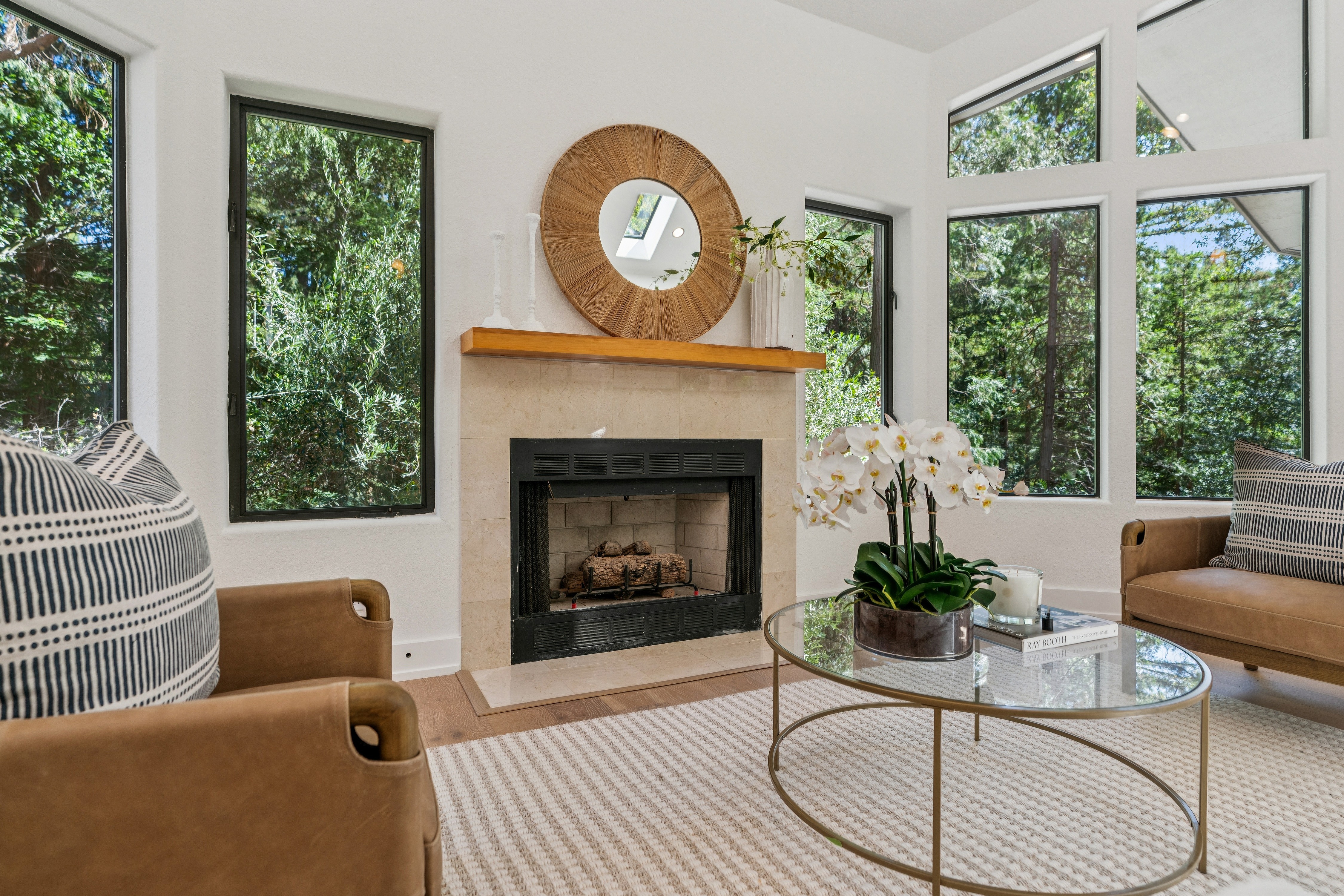 This bright and airy living room features a central fireplace with a light stone surround and a natural wood mantel, topped by a large circular woven mirror. Two leather armchairs with patterned accent pillows are positioned around a glass-topped coffee table, creating a cozy seating area. The space is framed by large windows that offer expansive views of the surrounding trees, filling the room with natural light.