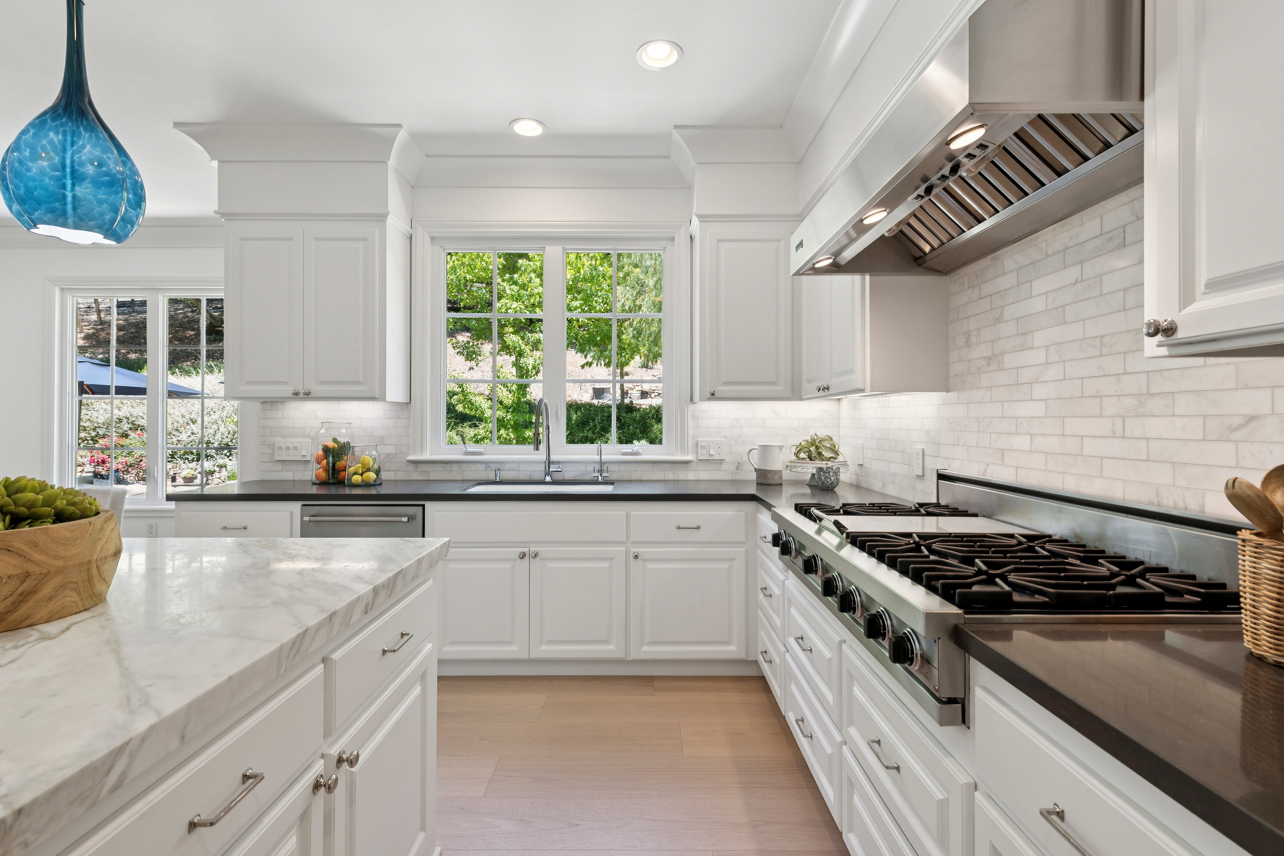 This bright and airy kitchen features crisp white cabinetry, a professional-grade stainless steel range with a matching hood, and a large central island topped with a light-toned stone surface. A subway-style backsplash runs along the walls, complemented by dark countertops and light wood-toned flooring. The perspective captures the functional layout, highlighting the sink positioned beneath a window that looks out onto lush greenery.