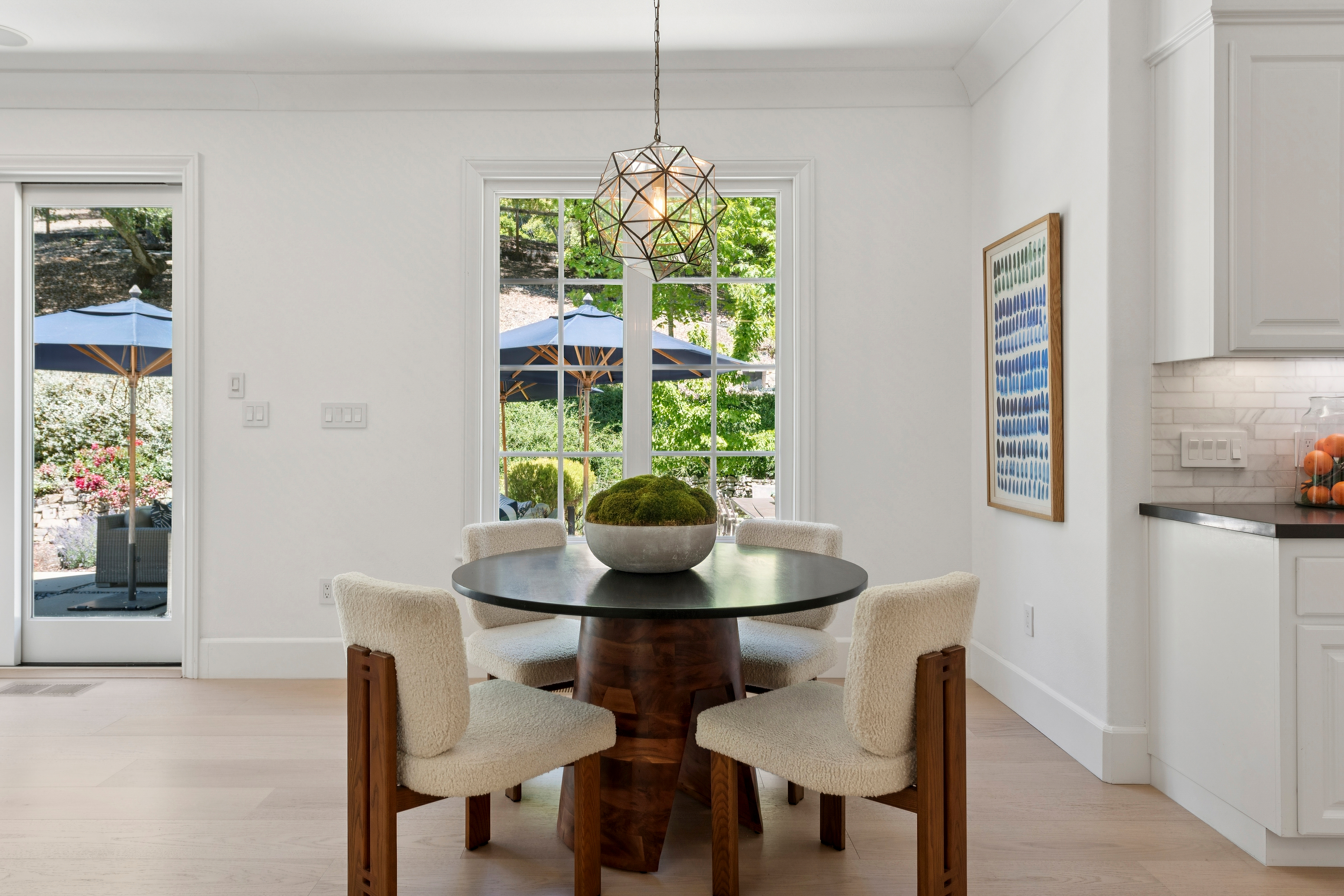 This bright and airy dining area features a round dark wood table paired with four cream-colored, textured upholstered chairs. A geometric, multi-faceted pendant light hangs above a central moss-filled bowl centerpiece, while large windows and a glass door provide ample natural light and views of the outdoor patio. The space is finished with light wood flooring and crisp white walls, creating a clean, modern aesthetic.
