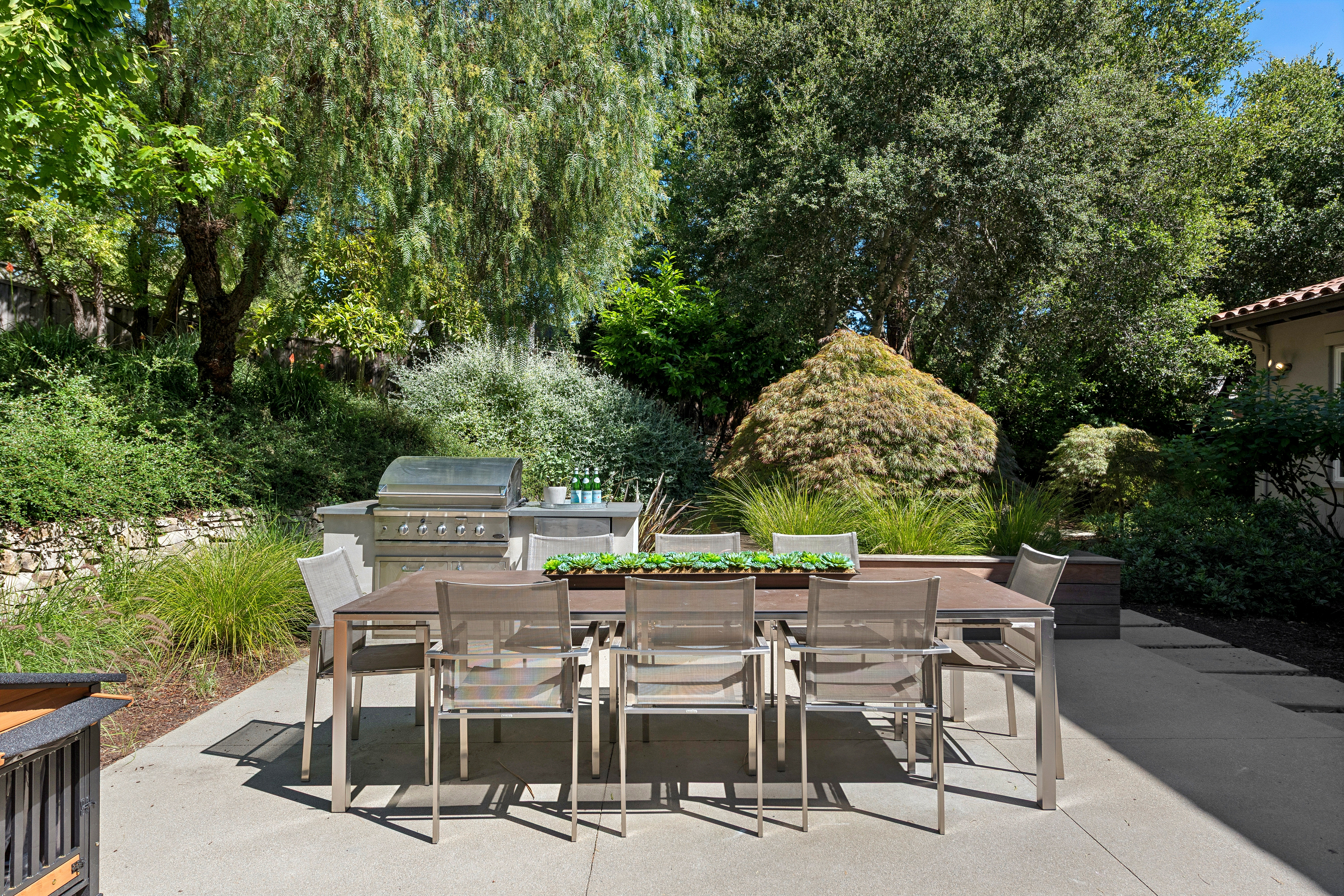 This outdoor patio space features a modern dining set with a long table and six mesh chairs, centered around a succulent planter centerpiece. Behind the dining area, a built-in stainless steel grill station is set against a lush, landscaped hillside filled with mature trees and greenery. The concrete patio provides a clean, functional space for al fresco dining, creating a serene and private backyard atmosphere.