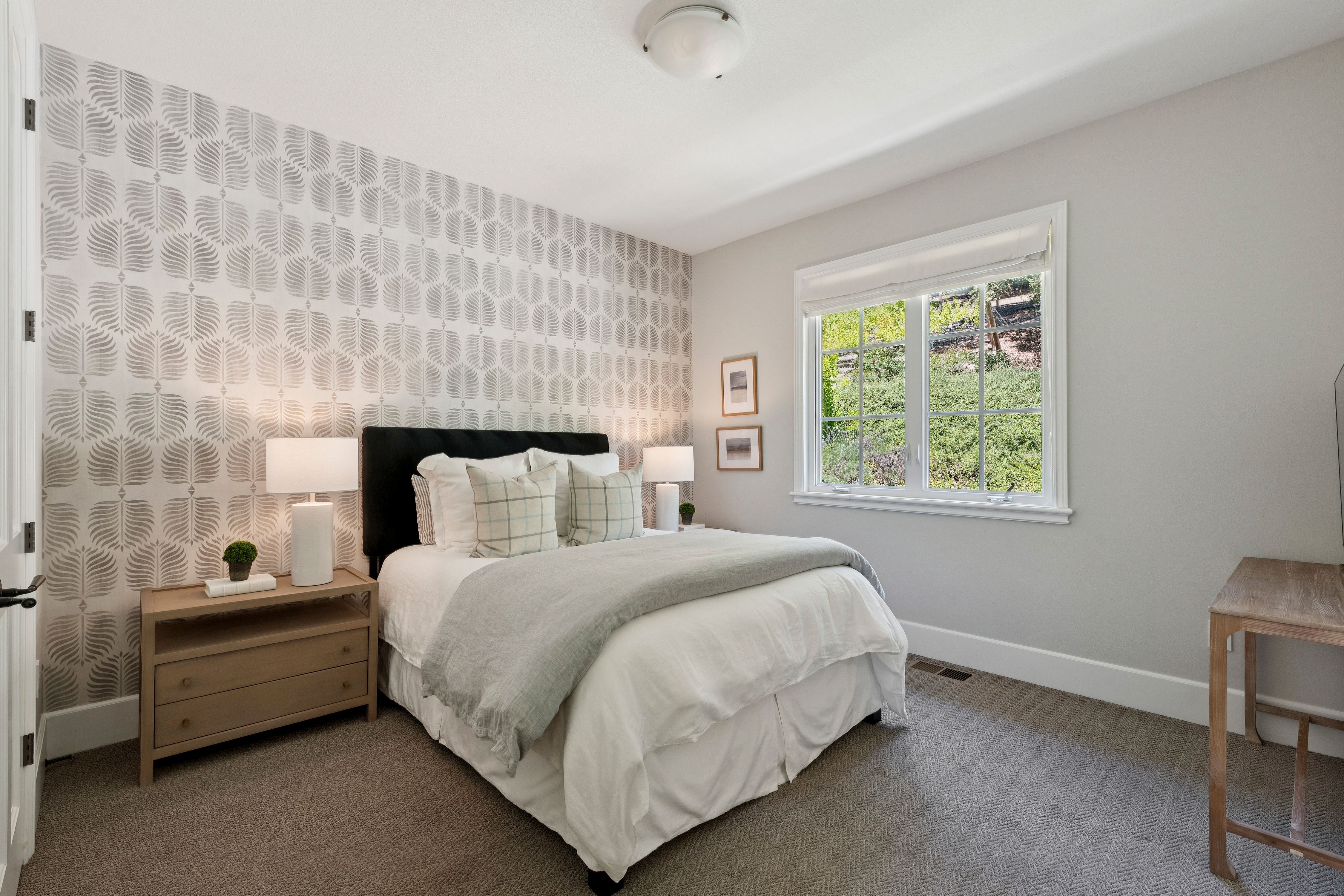 This cozy guest bedroom features a striking accent wall with a grey and white leaf-patterned wallpaper behind a dark-framed bed. The room is furnished with a light wood nightstand, a matching desk, and soft neutral bedding, creating a calm and inviting atmosphere. A large window allows natural light to fill the space, offering a view of the landscaped hillside outside.