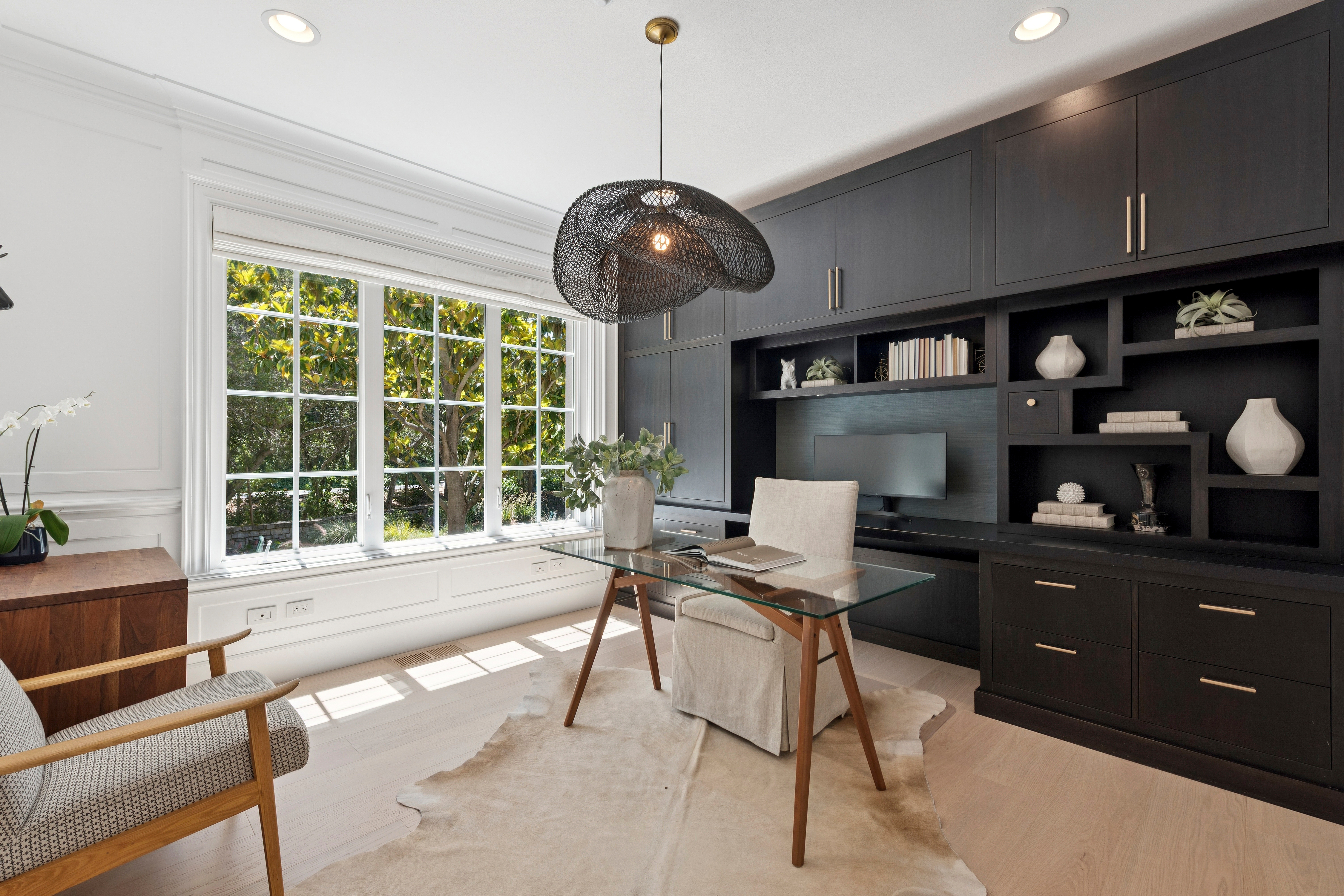 This sophisticated home office features a striking dark-toned built-in cabinetry unit with integrated shelving and a desk area. A glass-topped desk sits centrally on a light-colored hide rug, illuminated by a unique, sculptural pendant light fixture. Large windows provide ample natural light and a view of the lush greenery outside, creating a bright and productive workspace.